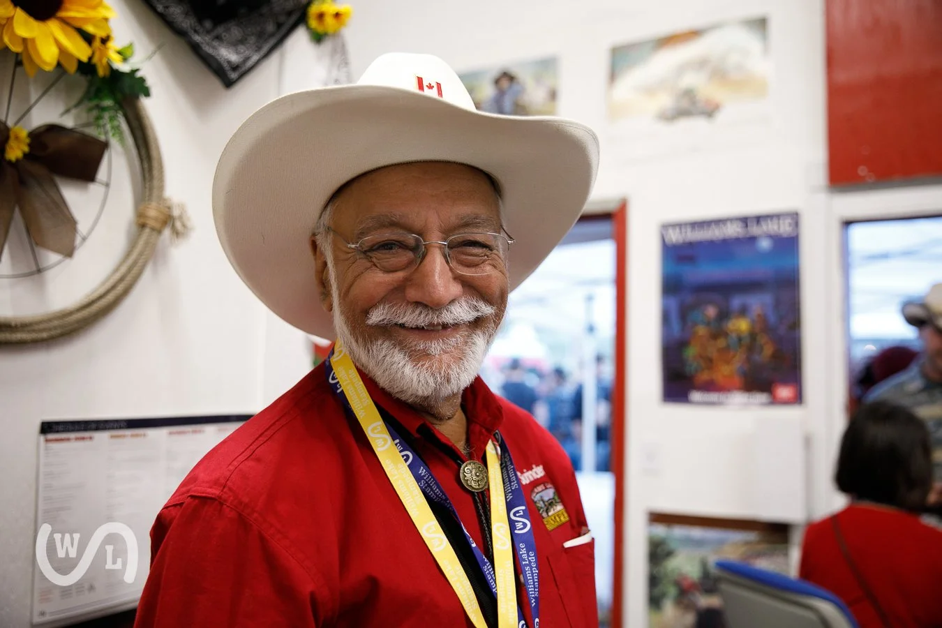 A smiling older man with grey hair and beard, wearing glasses, a cowboy hat with a Canadian flag, a red shirt, and a lanyard, inside a room with posters on the wall.