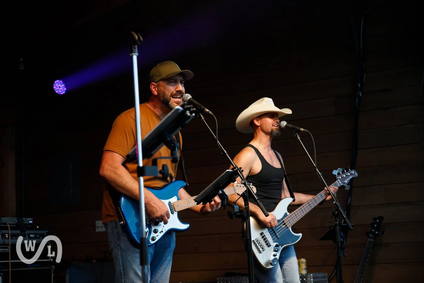 Two musicians performing on stage, one playing electric guitar and singing, the other playing bass guitar and singing, both wearing casual clothing and hats.