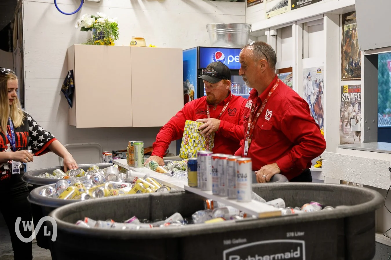 Two men in red shirts sorting cans of drinks at a recycling station in a room decorated with posters and a Pepsi logo.