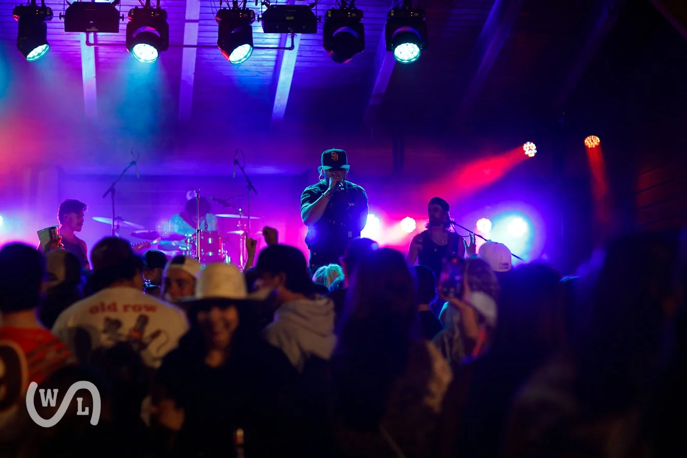 Live country music performance at a concert venue with colorful stage lighting, including a singer with a baseball cap, a drummer, and guitarists, surrounded by an audience.