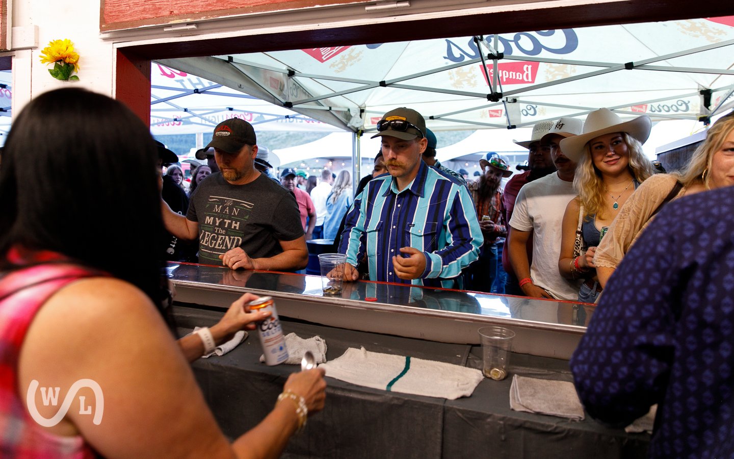 People lined up at a street fair or festival, waiting to purchase food or drinks from a vendor under a canopy, with some people holding cups and a woman handing a can to a customer.