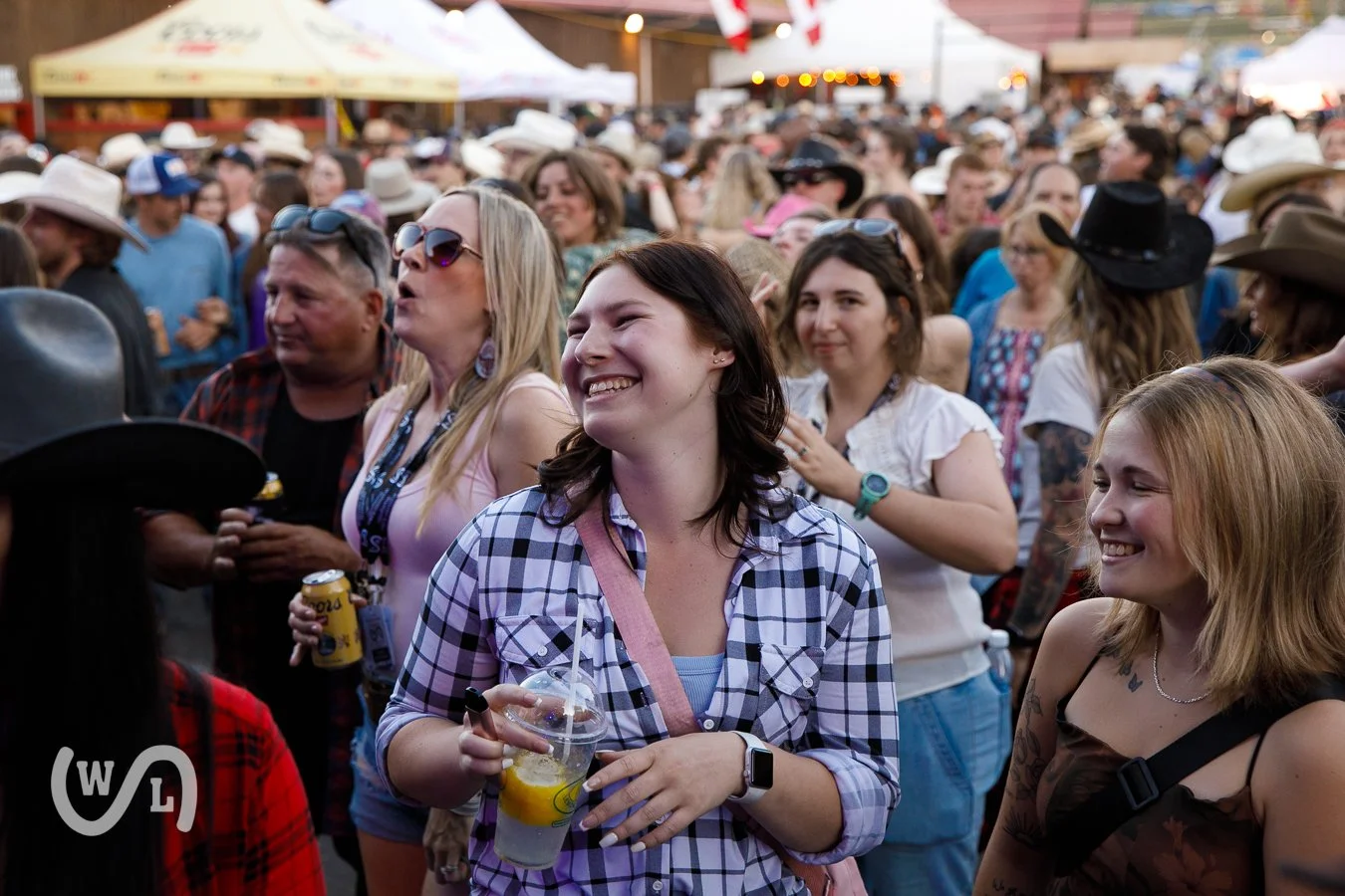 Crowd of people enjoying outdoor event, some wearing cowboy hats and sunglasses, with tents and flags in background, sunny weather.