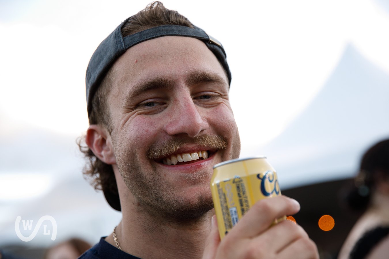 A young man with a mustache and a backward baseball cap smiling while holding a yellow can of Coors beer.