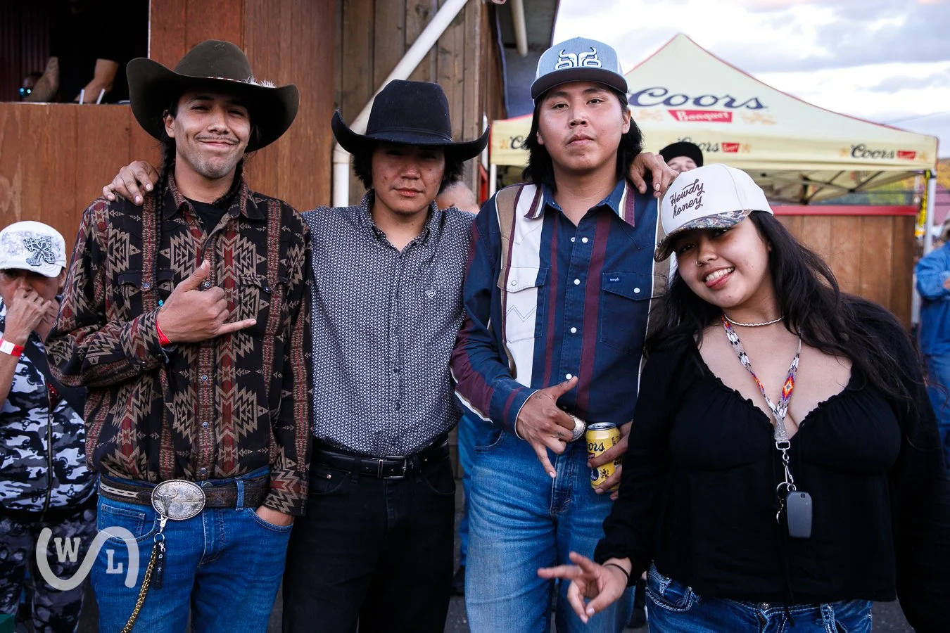Four people posing outdoors at a festival, with a yellow Coors tent in the background. Two men wearing cowboy hats and two women, one wearing a white hat and the other wearing a black hat. One man is giving a thumbs up, and they are all wearing casua