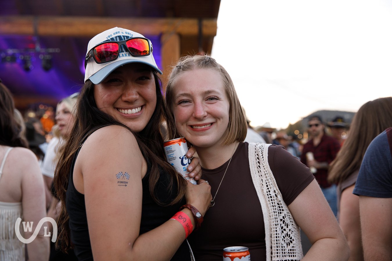 Two women smiling and hugging at an outdoor event, holding drinks, with a crowd in the background.