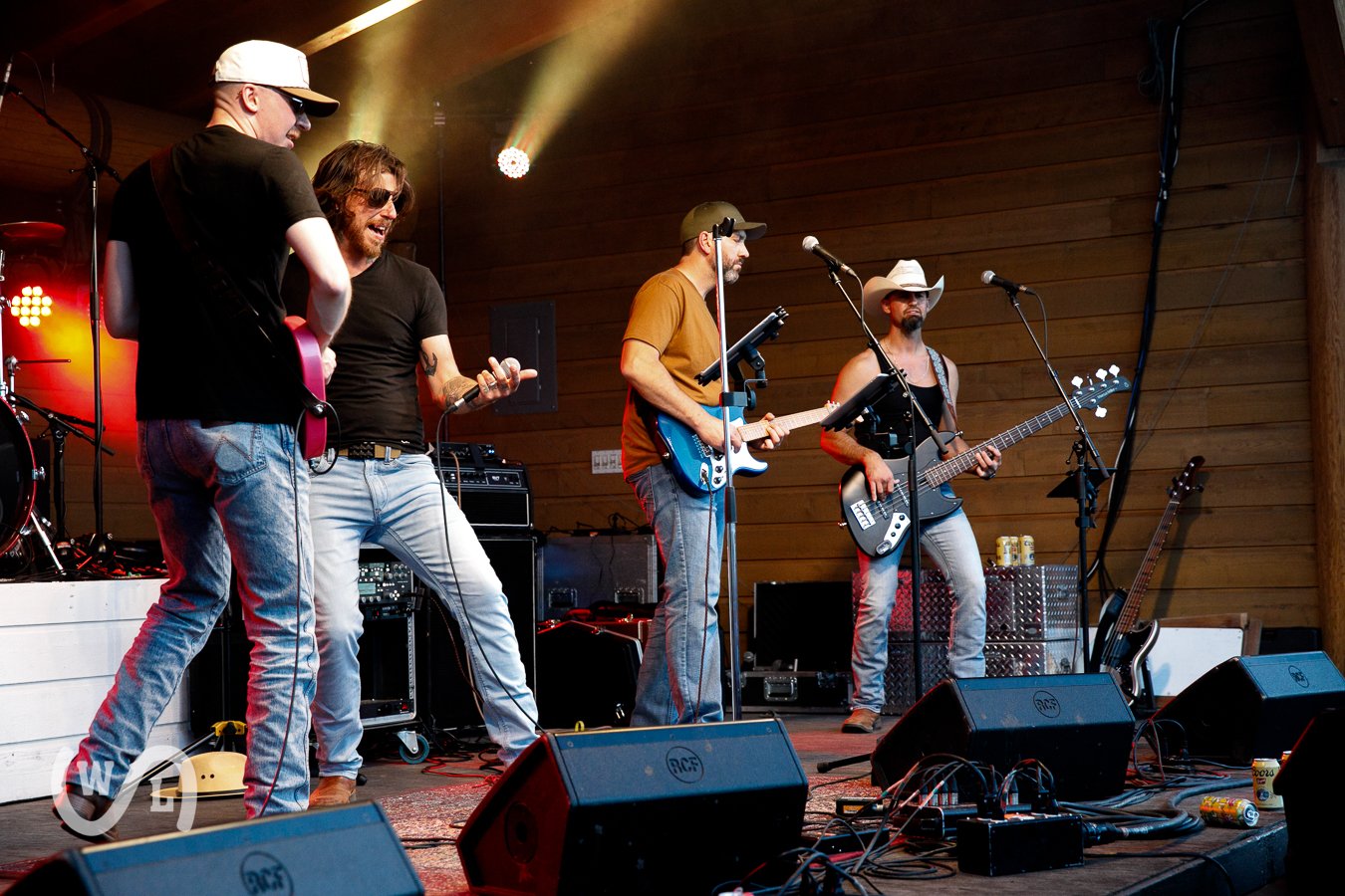 Four male musicians performing on stage with guitars, microphones, and audio equipment, in a wooden-walled venue with concert lighting.