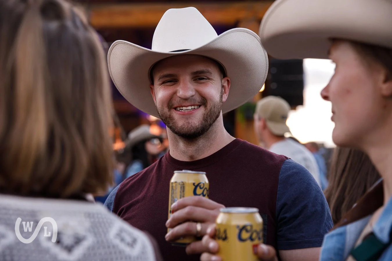 A man wearing a white cowboy hat and maroon and navy shirt smiling while holding a Coors Light beer can, standing and talking with two women also wearing cowboy hats at an outdoor event.