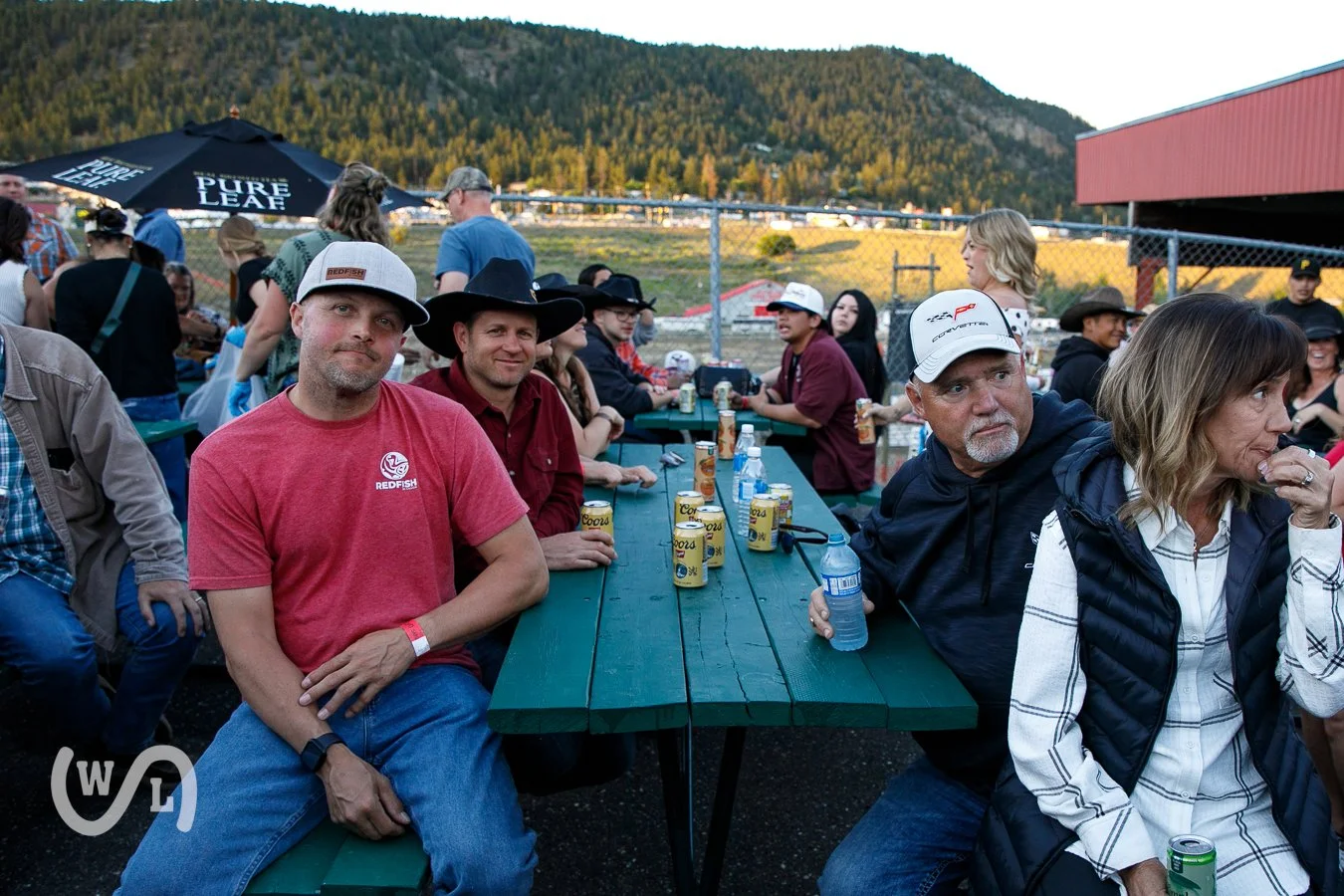 People sitting at a long green picnic table outdoors, with beverages in cans and bottles, enjoying a social gathering or event in a rural setting with mountains in the background.