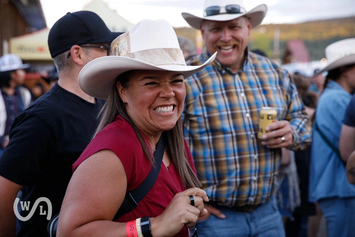 A woman wearing a white cowboy hat and a red shirt is smiling and appears to be enjoying herself at an outdoor event, with a man in a plaid shirt holding a drink nearby.
