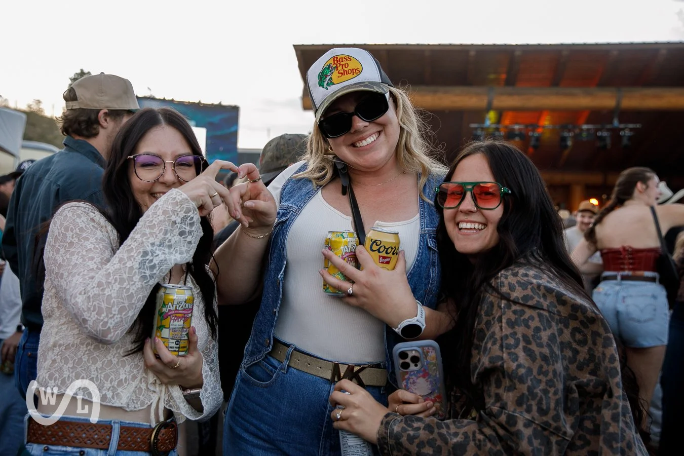Three women smiling at an outdoor event, holding canned drinks, with a stage and crowd in the background.