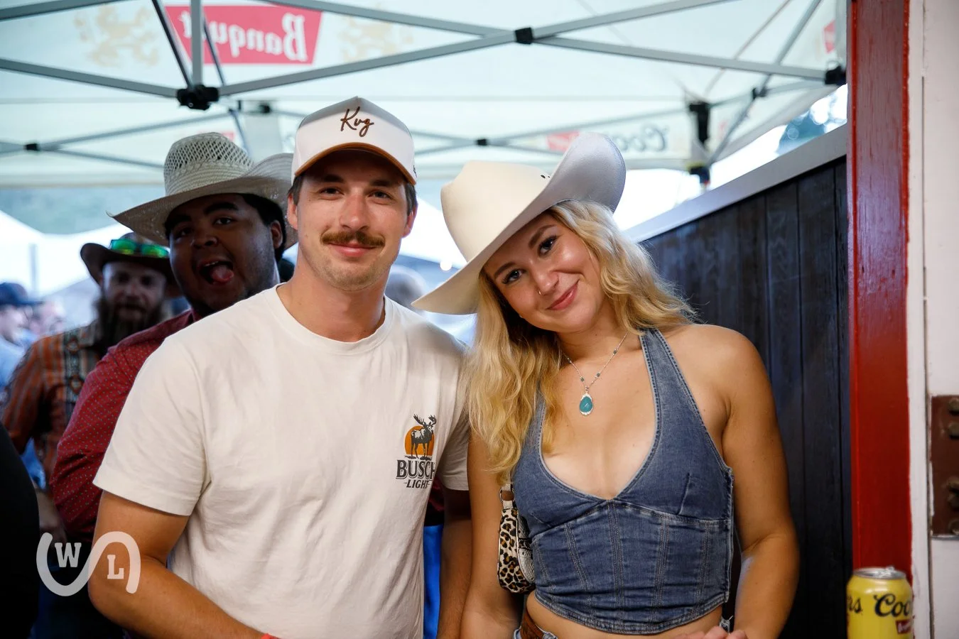 Group of people at an outdoor event, with a man and woman in the foreground wearing cowboy hats, smiling and posing for the camera. The man has a mustache and wears a white t-shirt, while the woman has long blonde hair and wears a denim sleeveless to