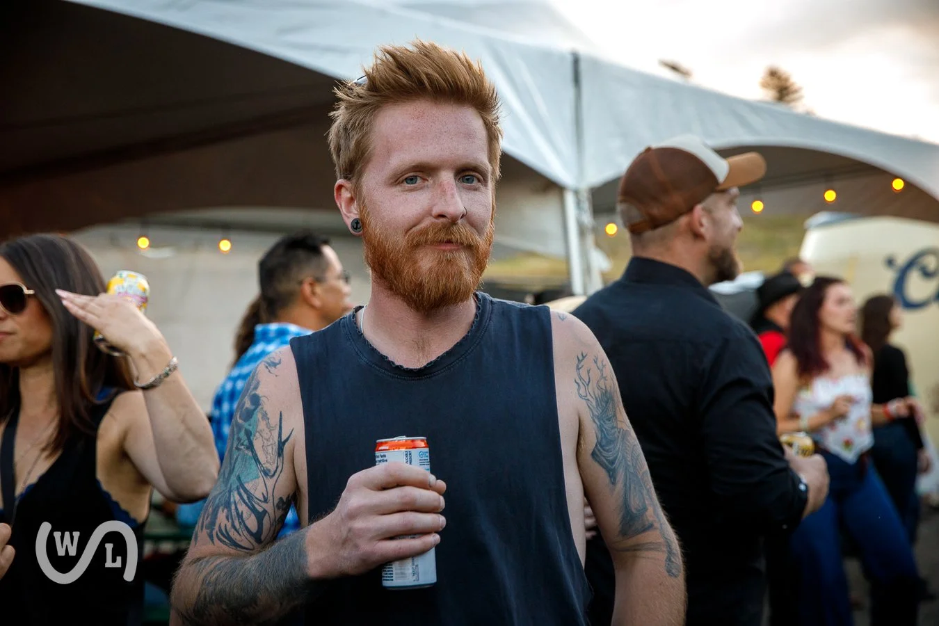 A red-haired man with a beard and tattoos on his arms holding a can of beverage at an outdoor gathering during sunset, with tents and other people in the background.