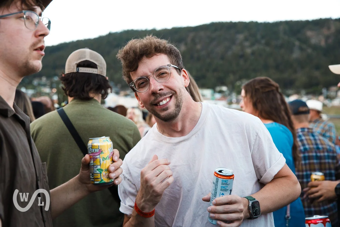 Group of people socializing at an outdoor event, with a mountain in the background, drinking cans of soda, smiling and enjoying themselves.