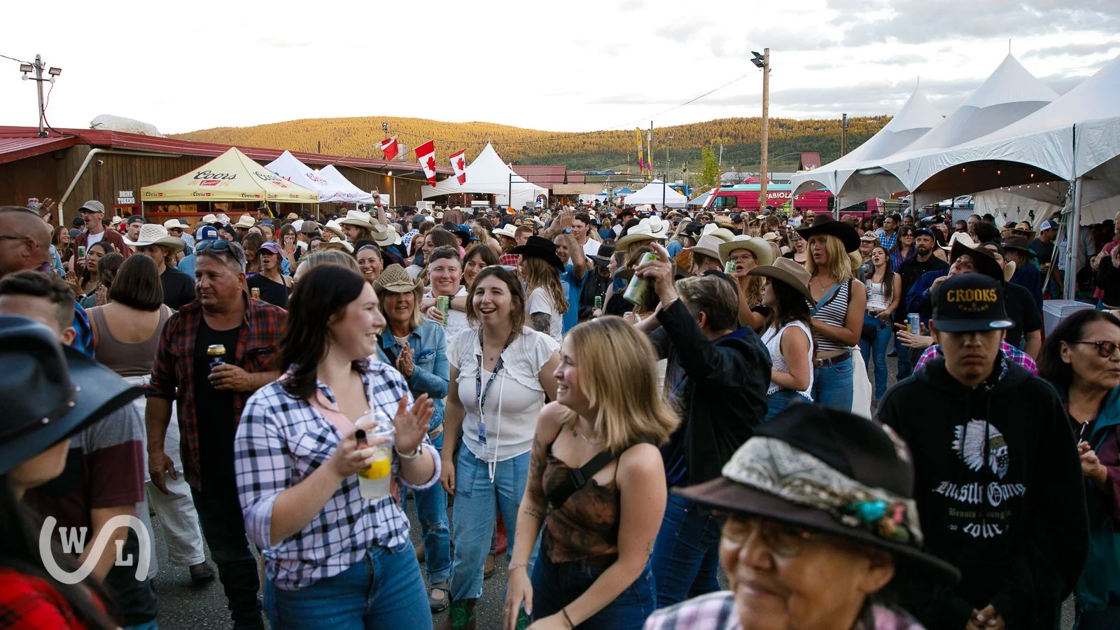 Crowd of people at an outdoor country music festival, many wearing cowboy hats and holding drinks, with tents and flags in the background.