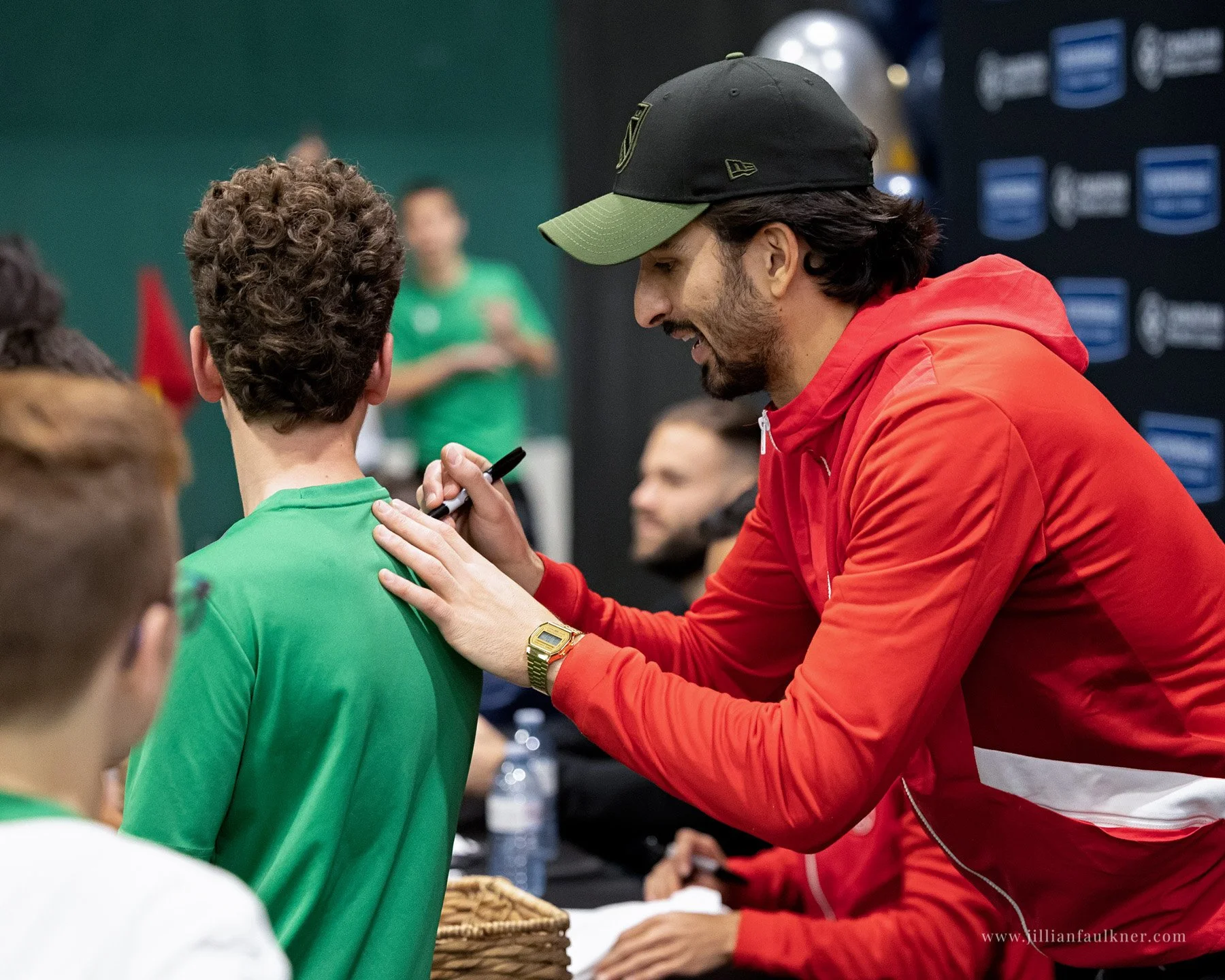 Soccer athlete candid photo taken during the soccer event and photographed by Calgary portrait and event photographer. The athlete is in red hoodie signing autograph on boy's green shirt at event.