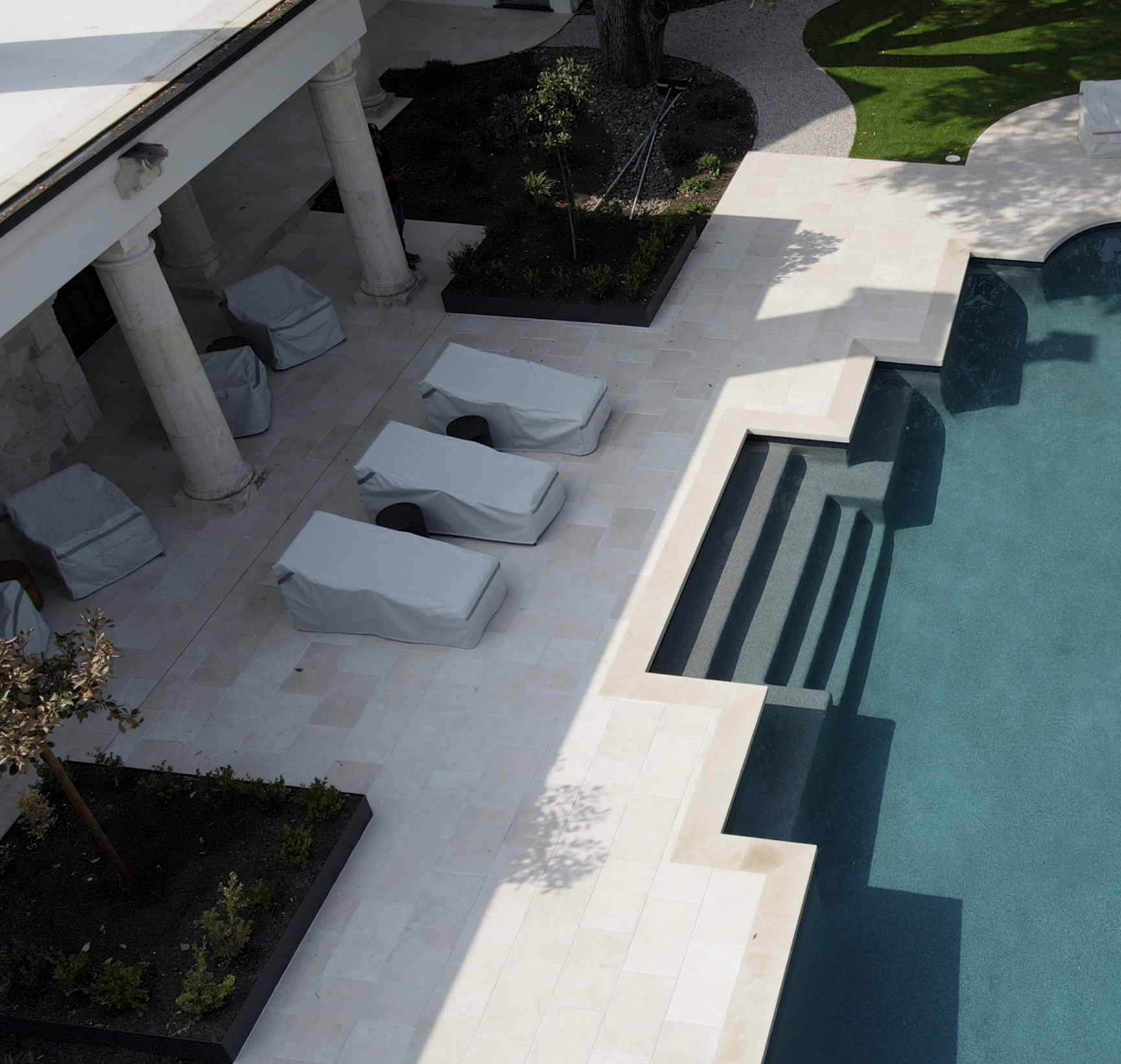An outdoor seating area with a wooden bench, gray cushions, and colorful pillows. A rectangular fire feature with flames is in front, surrounded by a concrete base. Behind are lush green tropical plants and a wooden fence.