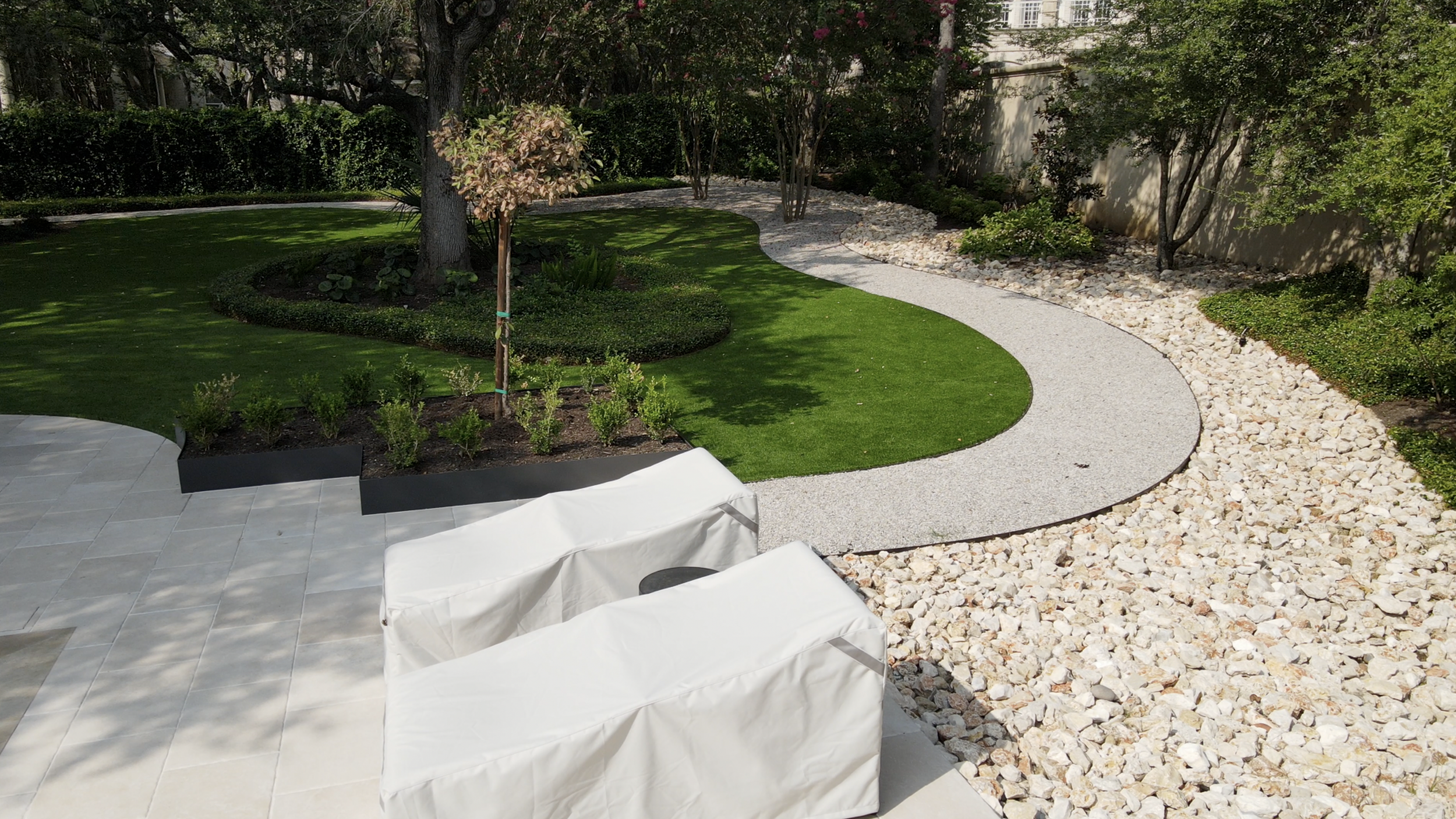 Outdoor patio area with a stainless steel grill, bar seating with four chairs, and a white table. There's a garden with various plants and succulents, a green artificial grass surface, and a backdrop of tall bamboo plants.