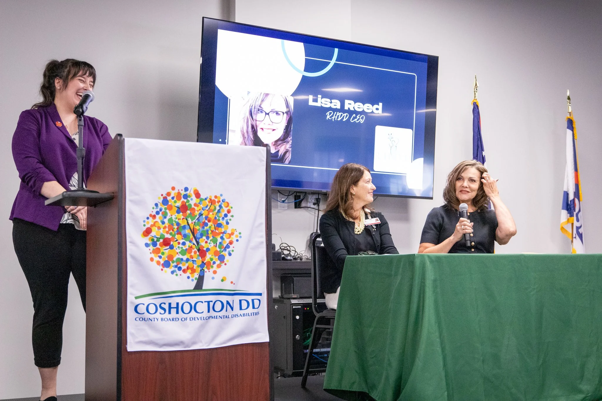 image of three women on a stage.  woman on the left is laughing, standing behind a podium.  two women on the right are seated behind a table.  Woman on the right is speaking into a microphone.