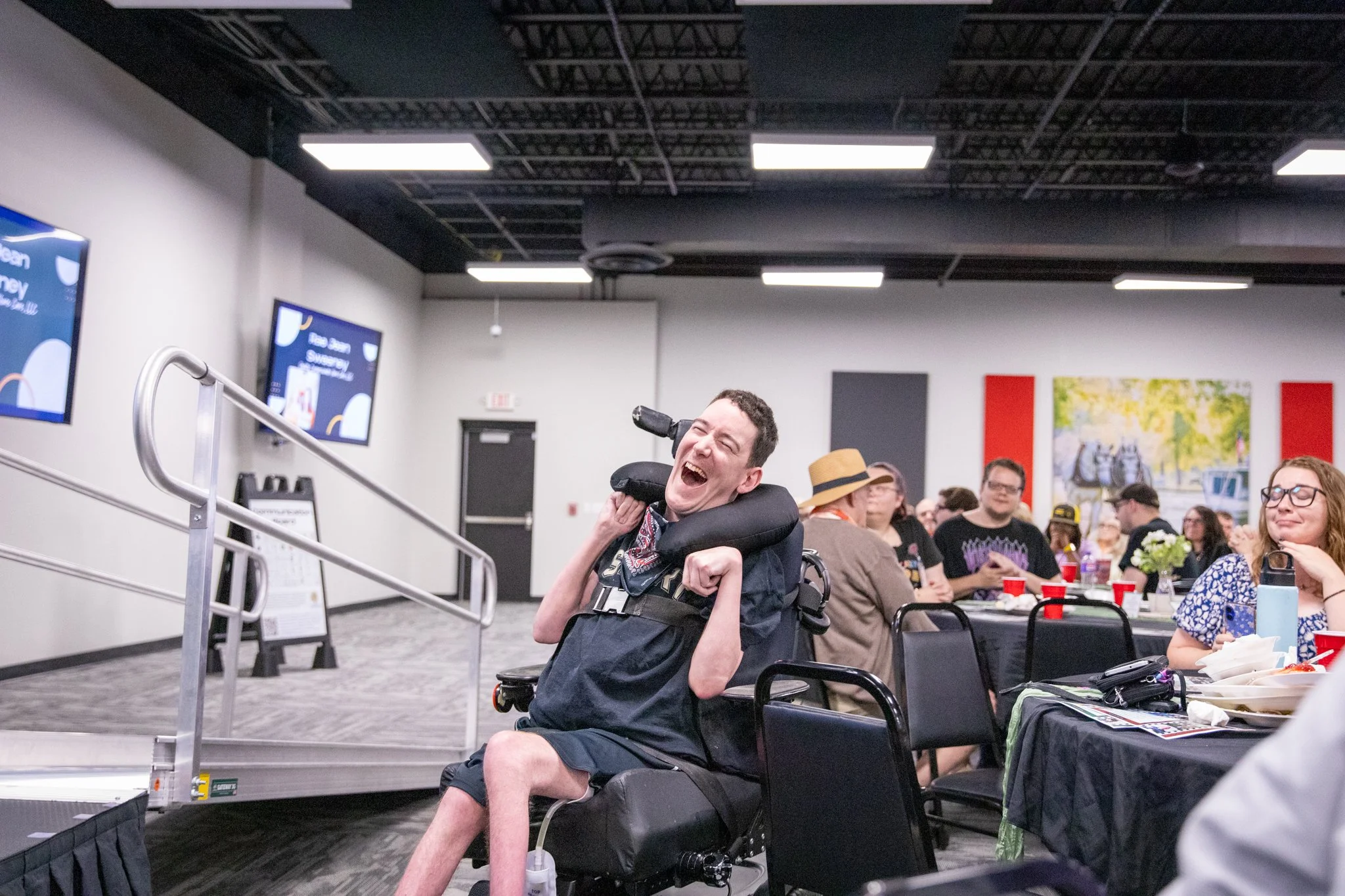 image of people sitting in the audience -- special highlight on a person in a wheelchair laughing and smiling while watching people on stage