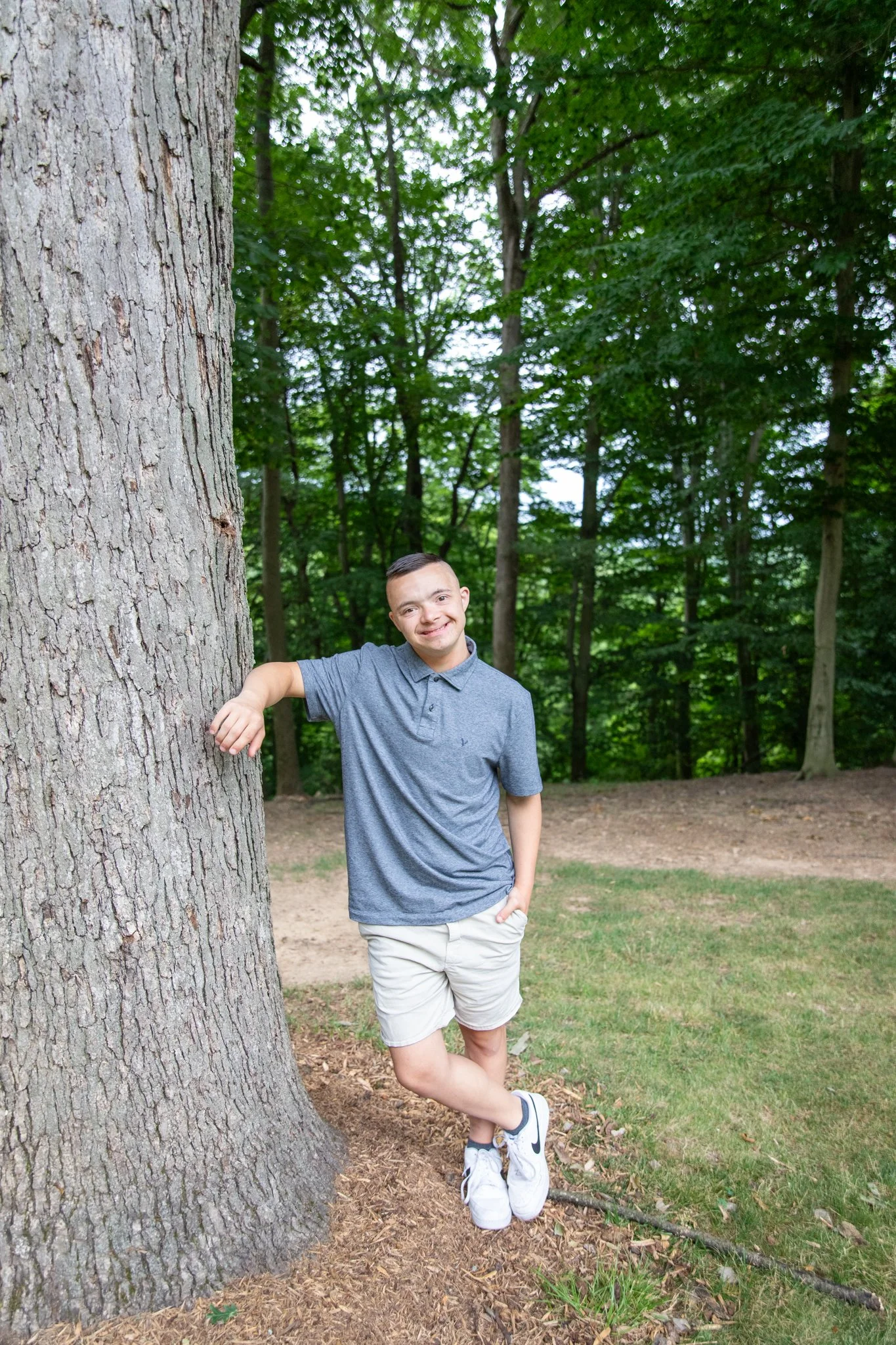 image of a young man with down syndrome wearing a grey polo smiling at the camera, leaning against a tree.  there are many green pine trees in the background.