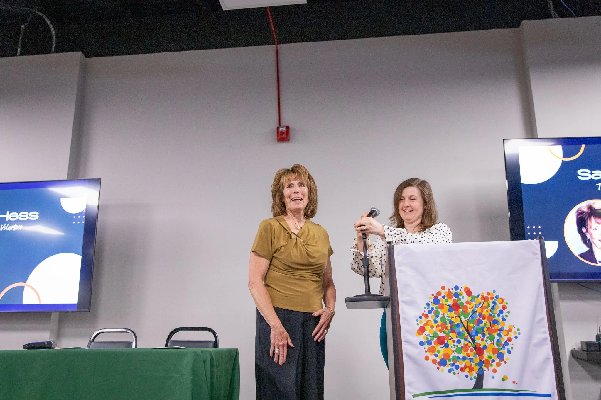 image of two women on a stage.  woman on left is smiling and looking at the audience.  woman on the left is putting a microphone on a stand, smiling.
