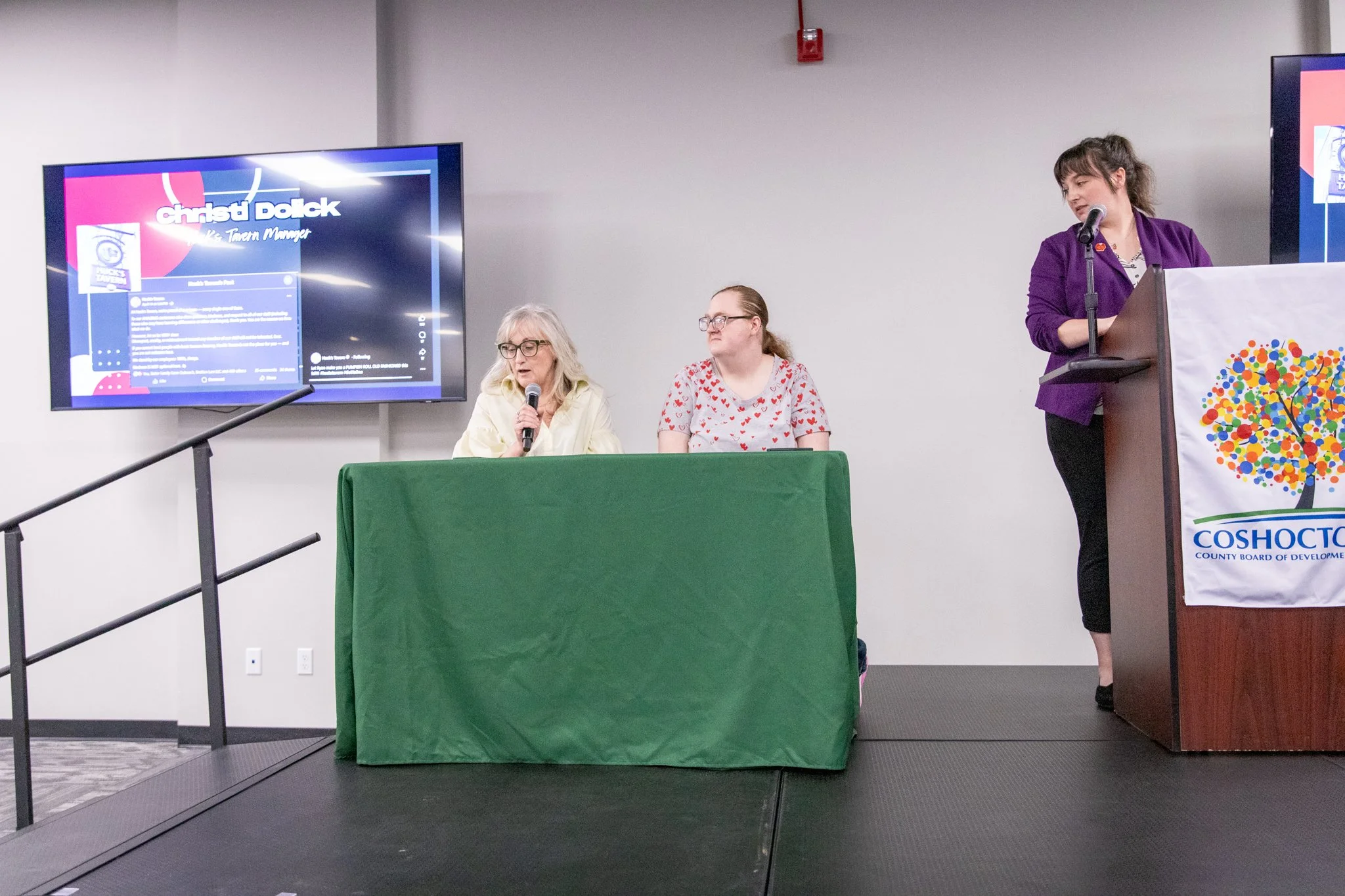 image of three women on a stage.  Two women on the left are seated behind a table, third is standing at a podium.  Woman on the far left is speaking into a microphone.