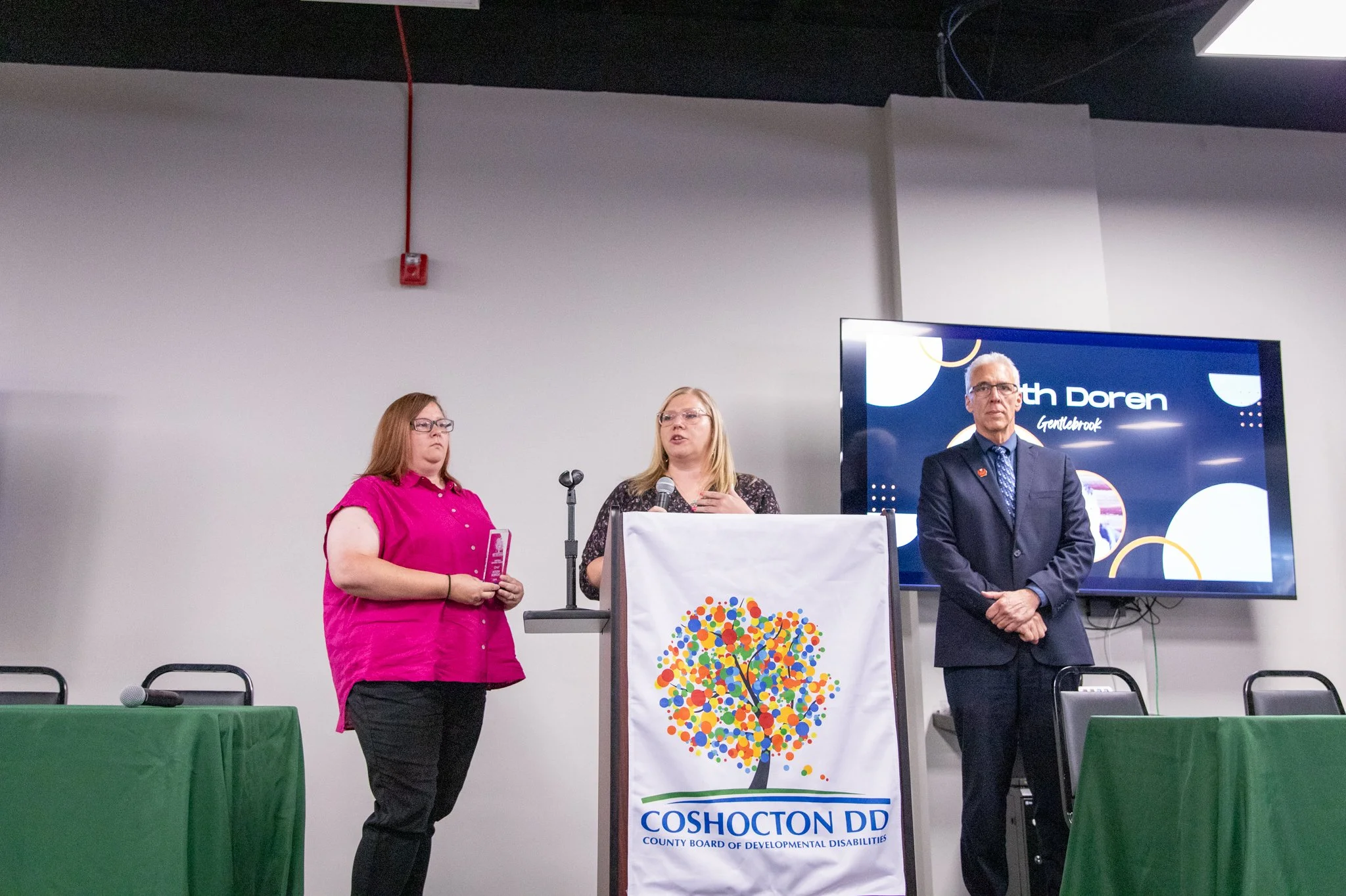 image of three people standing on a stage.  woman on the left is holding an award.  woman in the middle is speaking into a microphone, accepting an award on behalf of her father.  man on the right is looking at the crowd.