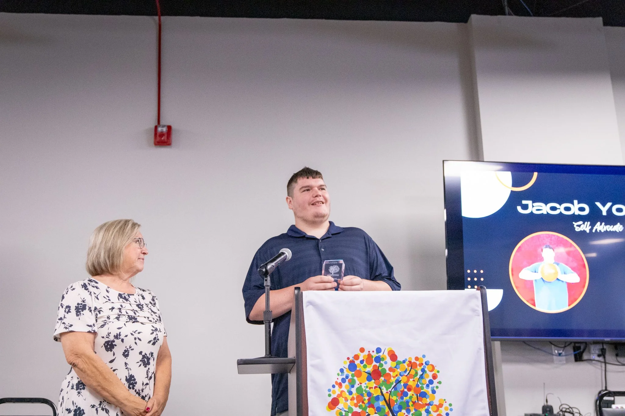 image of two people standing on a stage.  woman smiling and looking at man to her right.  man is smiling and speaking into microphone.