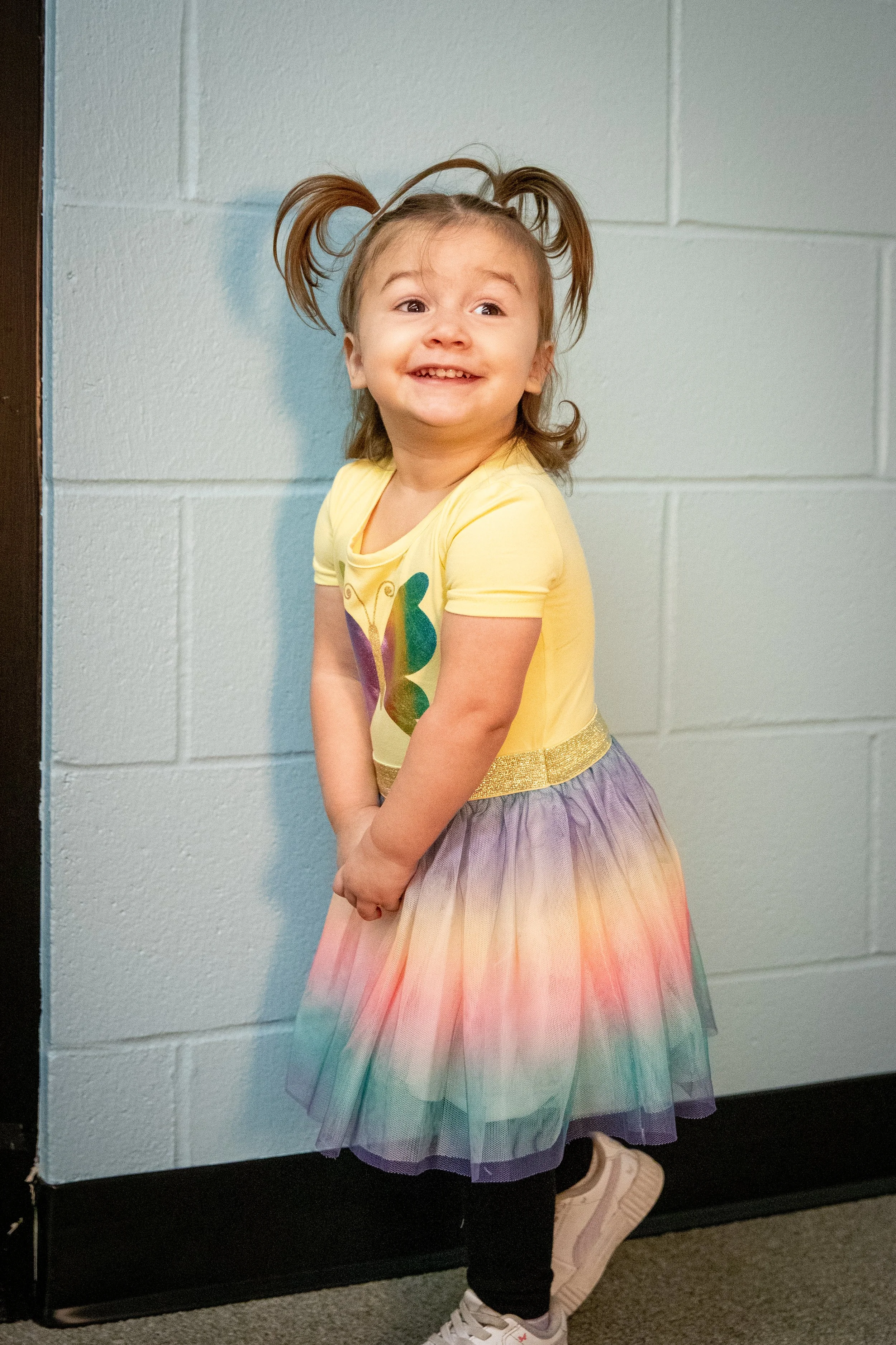 vertical image of young girl with brown hair in pig tails smiling.  she is wearing a yellow shirt with a rainbow butterfly and a rainbow skirt