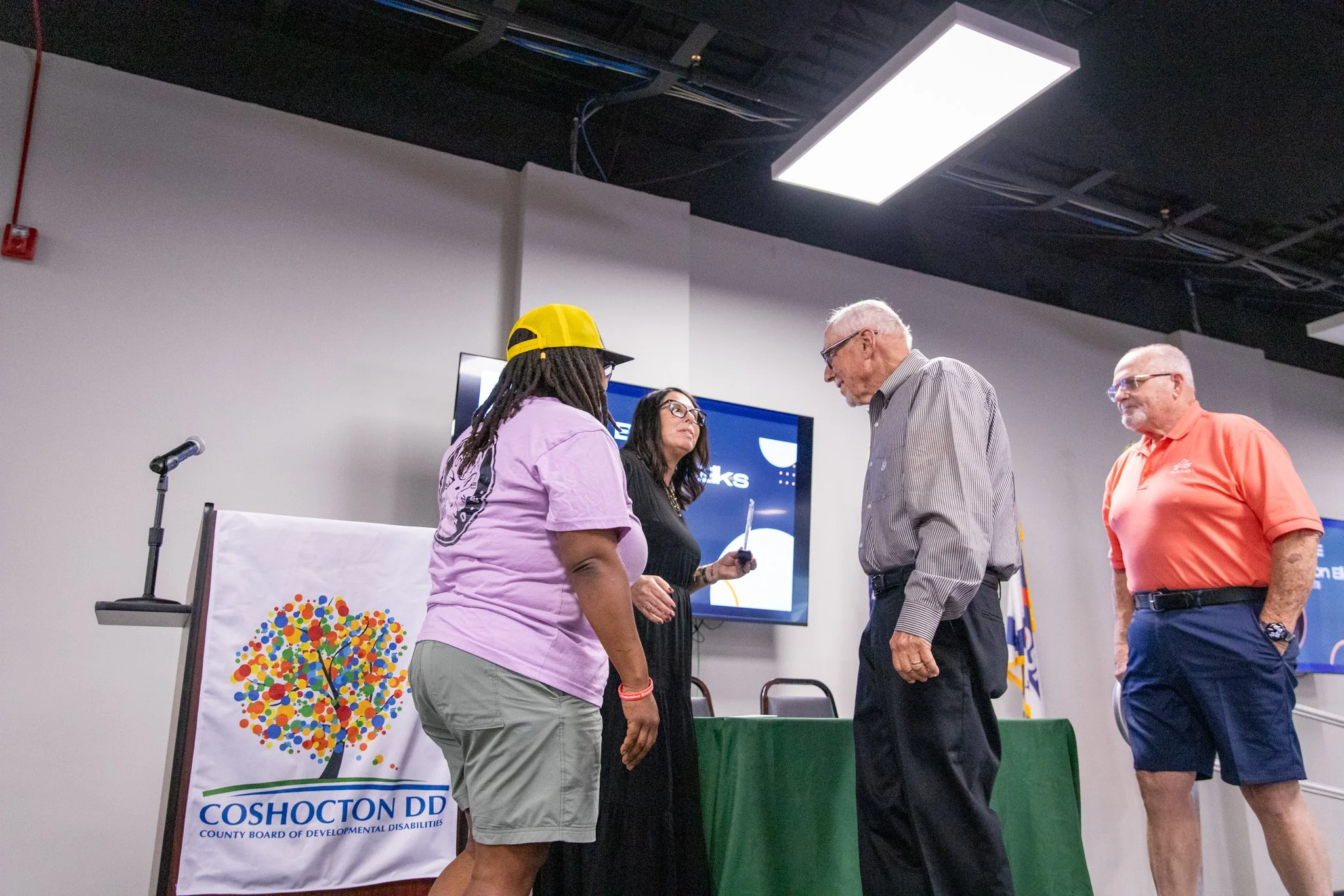 image of 4 people on a stage.  woman on the left is looking at the other three people.  woman in black dress is holding an award and looking at the man on her left.  First man is looking at woman in black dress, man in orange is looking at the group.