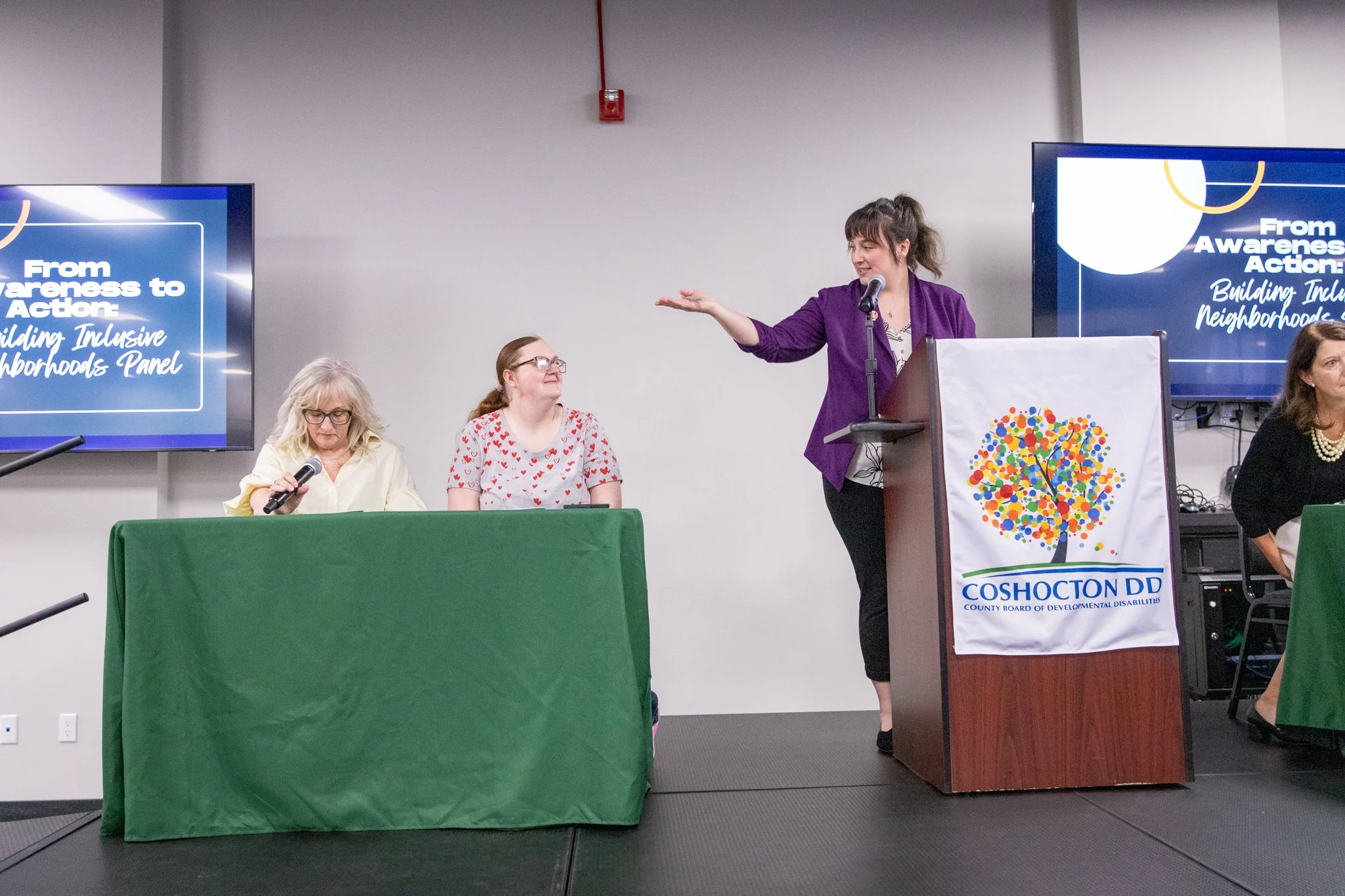 image of people on a stage.  two woman on the left are seated behind a table.  Woman in the middle is standing behind a podium gesturing to those two seated women.