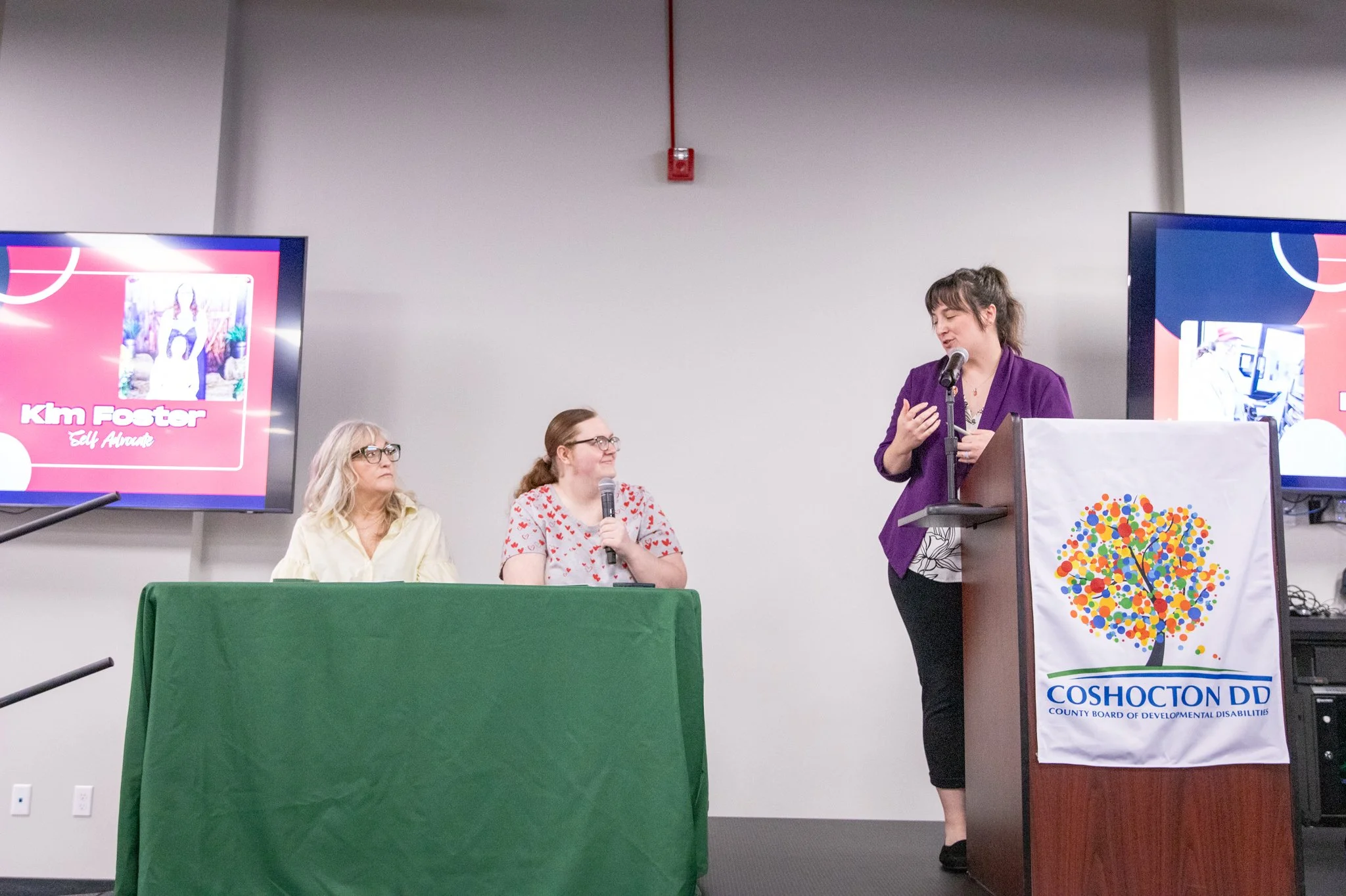 image of three women on a stage.  Two women seated behind a table, third is standing behind a podium.  Podium woman is speaking into a microphone and looking at those two women by table.