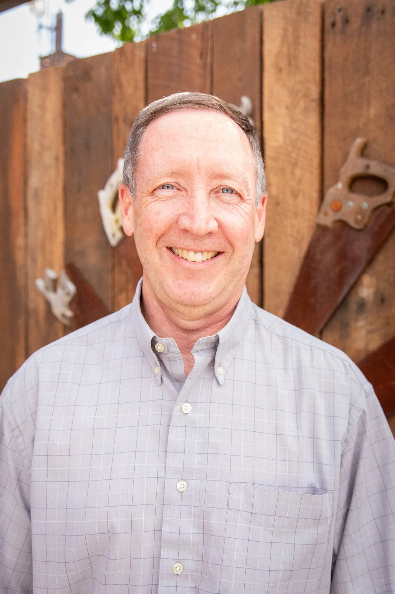 Close-up outdoor portrait of a person with short hair, wearing a light gray button-up shirt. The person is standing in front of a wooden fence decorated with metal hand tools, with greenery visible above and a softly blurred background.