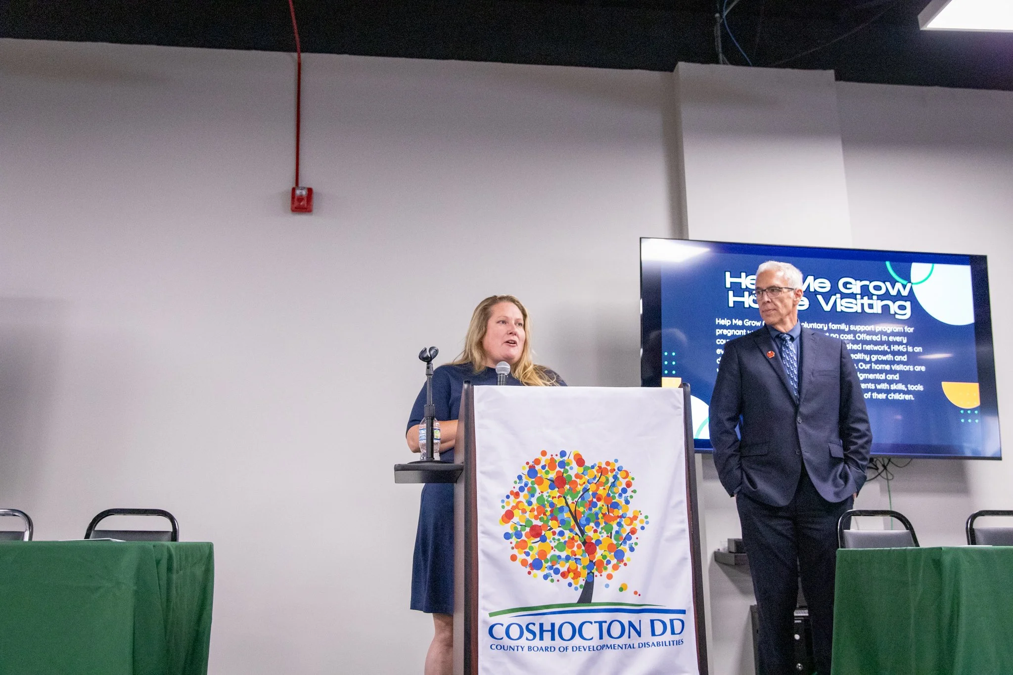 photo of two people standing on a stage. the woman is behind a podium speaking into a microphone.  man is standing off to the side looking at her.