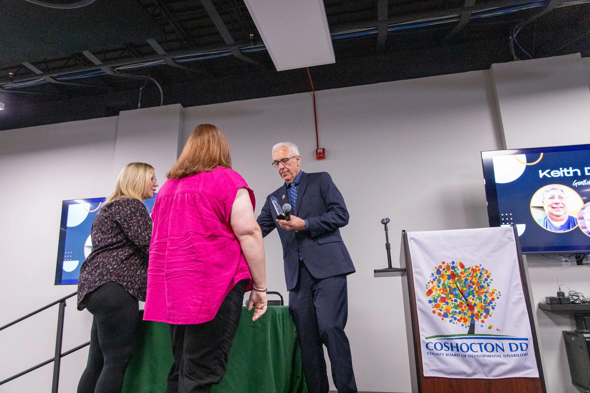 image of three people on a stage.  two women on the left are looking at the man on the right.  Man is holding an award and presenting it to the two women.