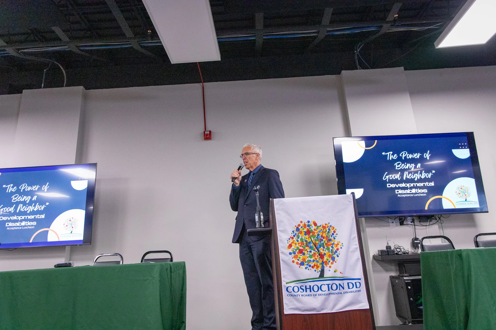 photo of man with white hair on a stage, holding a microphone speaking to audience.  Coshocton DD logo on podium.
