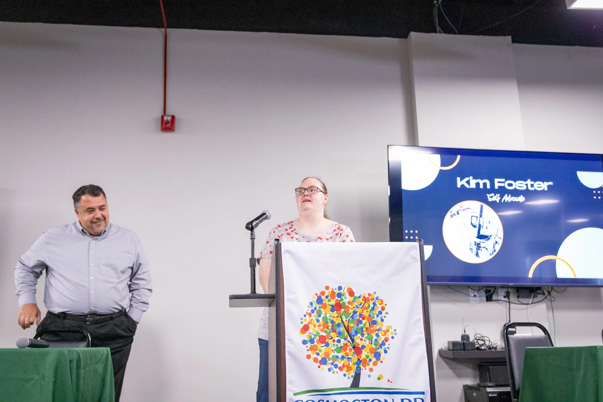 image of two people standing on a stage.  man on left smiling, woman on the right speaking into a microphone.