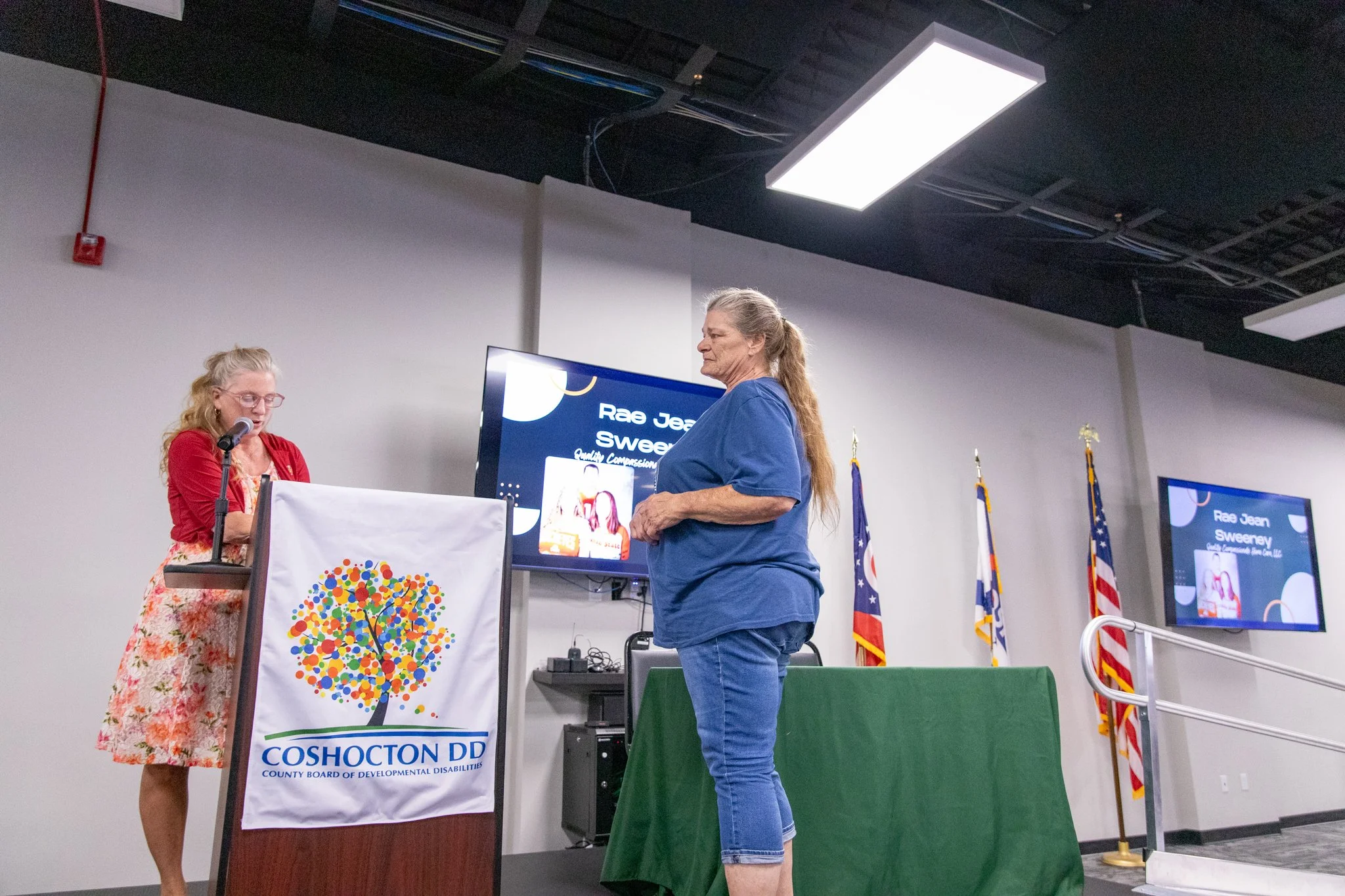 image of two women on a stage.  woman on left is speaking at a microphone, woman on the left is watching the woman on the left.