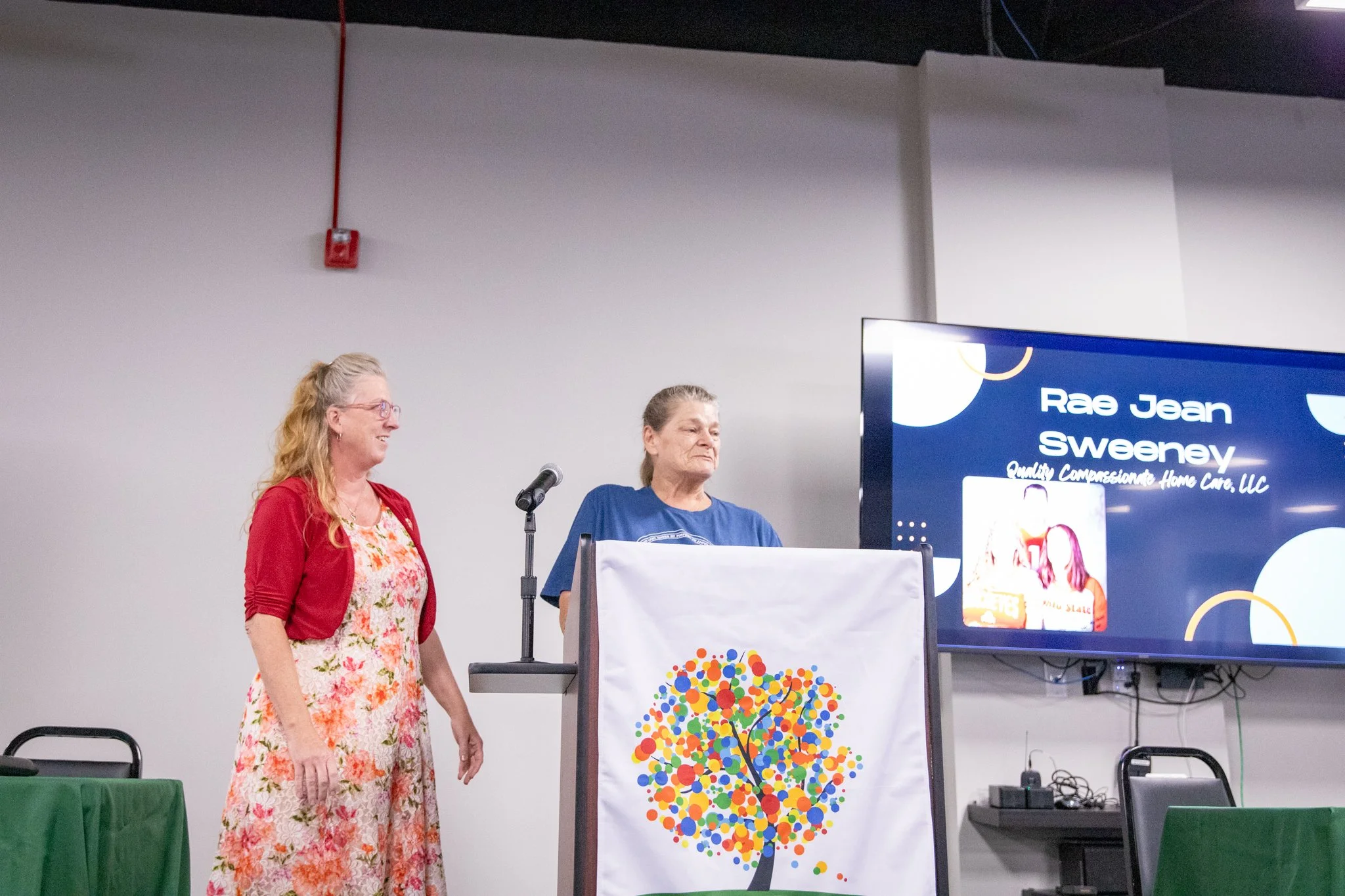 image of two women standing on a stage.  woman on left is smiling and looking at woman on right.  woman on right is behind the podium and looking out at the crowd.