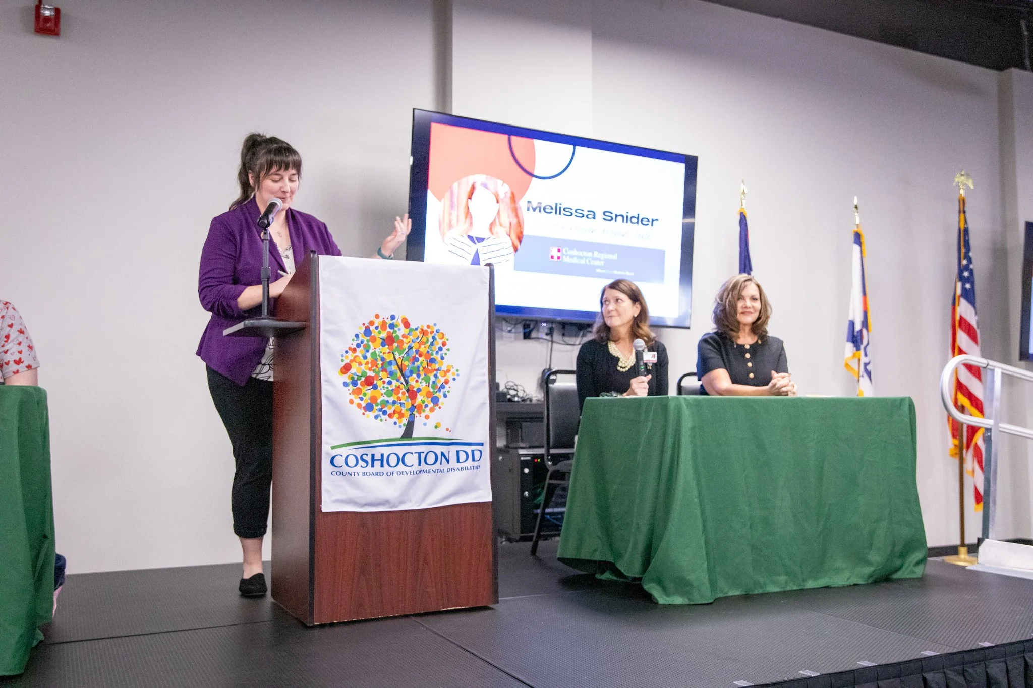 image of three women on a stage.  woman on the left is standing behind a podium.  the other two are to the right seated behind a table.