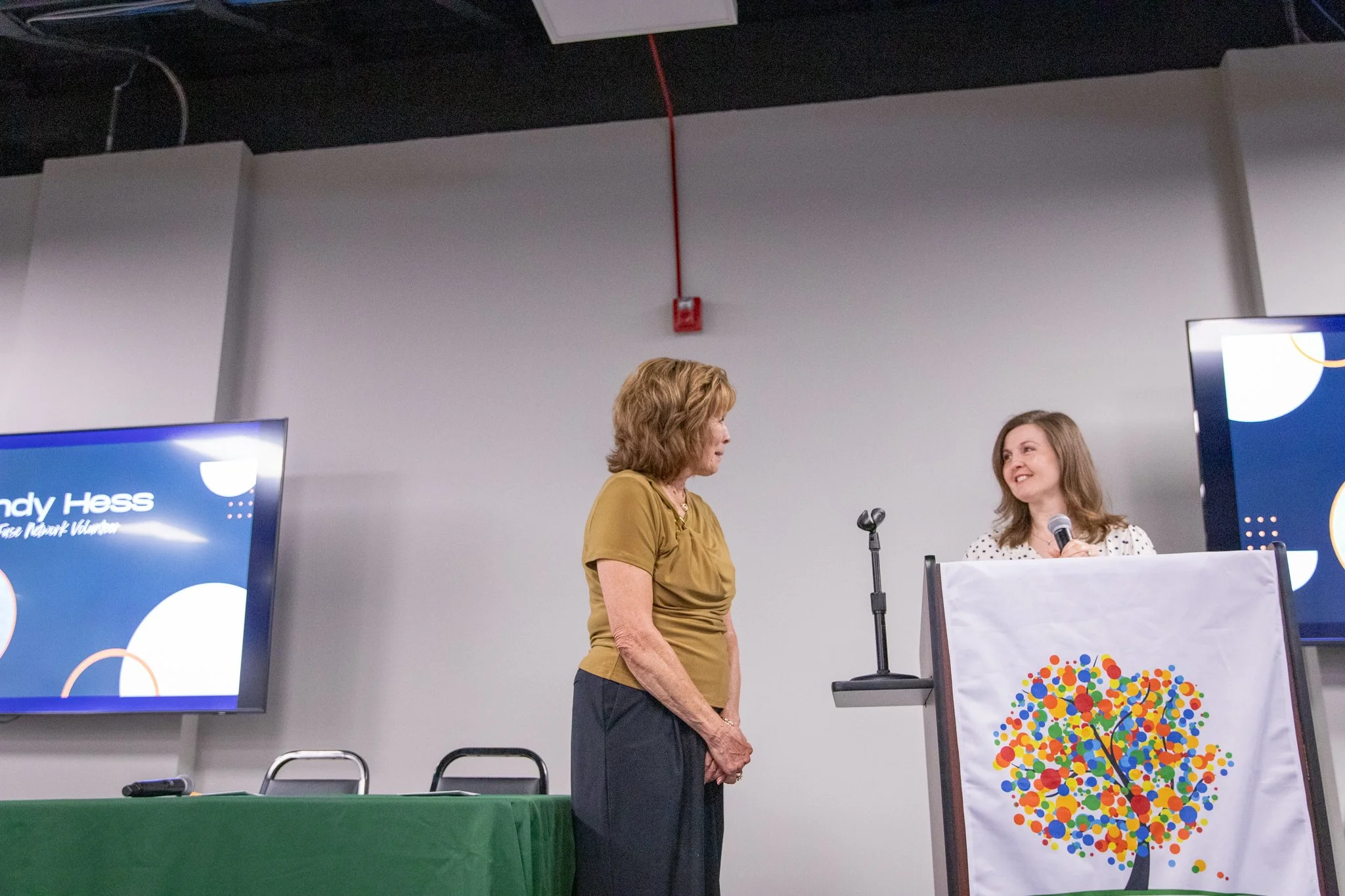 photo of two women standing on a stage.  woman on the left is looking at the woman on the right,  woman on the right is speaking into a microphone and looking happily at the woman on the left.