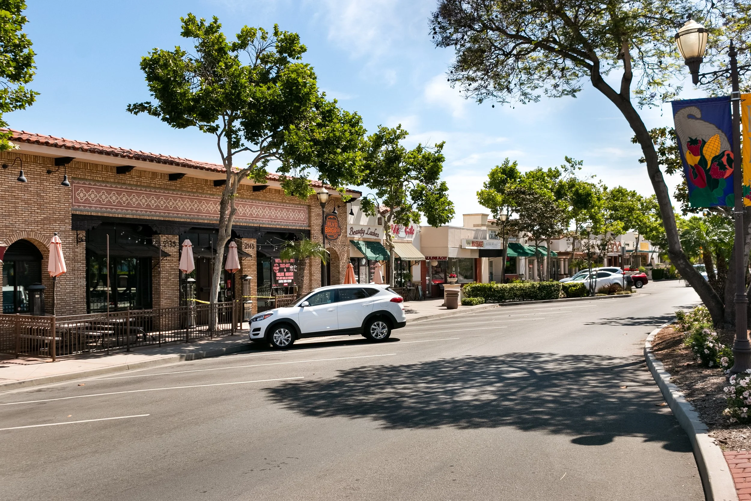 A street view in a shopping area with trees, storefronts, and parked cars, including a white SUV. The storefronts have outdoor seating and signage for various businesses.