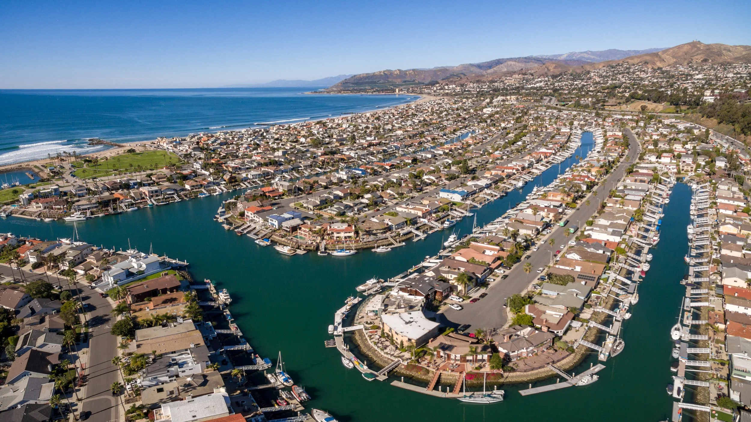 An aerial view of a coastal residential area with a marina filled with boats, facing the ocean with a beach, and mountains in the background.