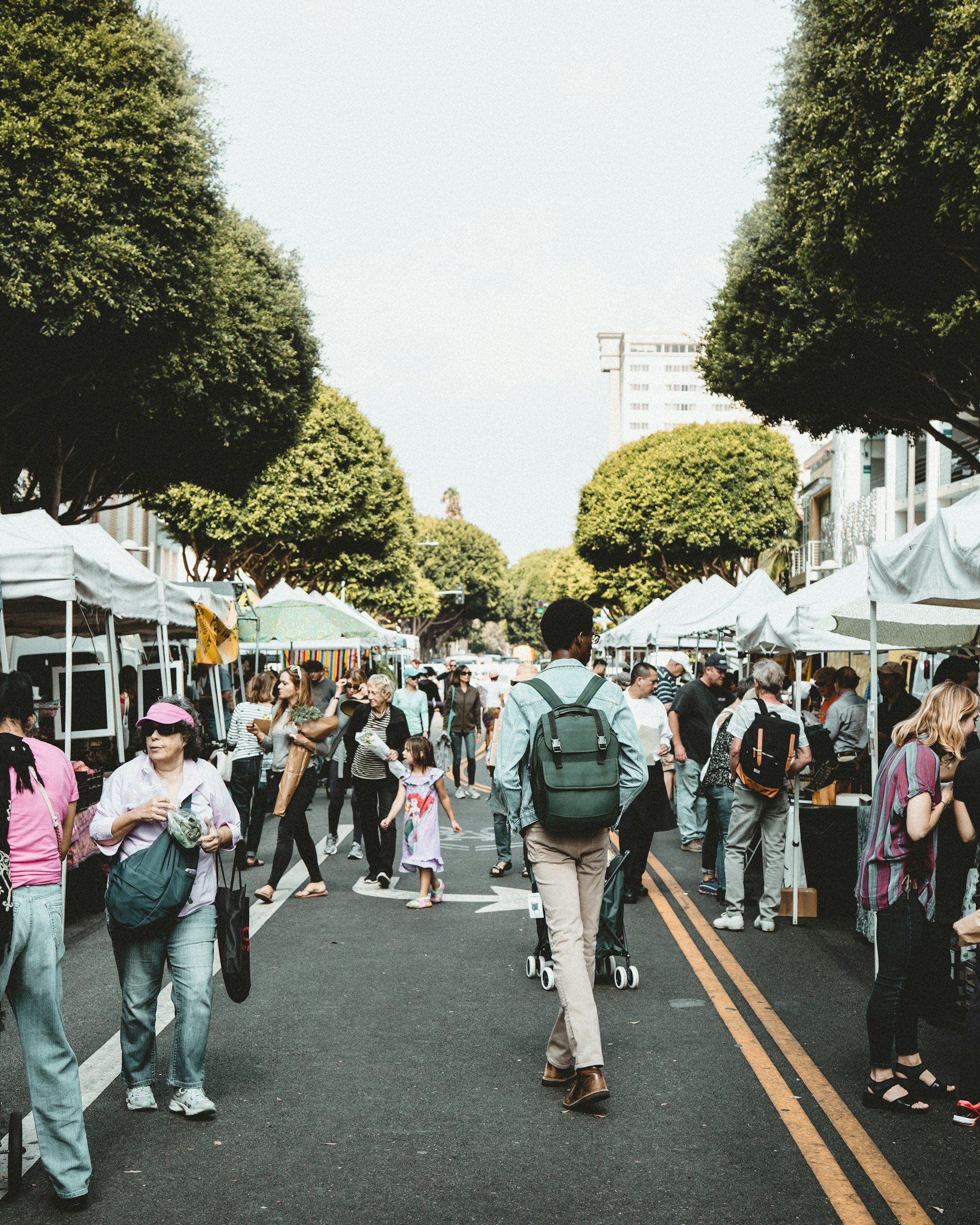 People walking and shopping at an outdoor street fair with white tents on both sides, trees overhead, and a tall building in the background.