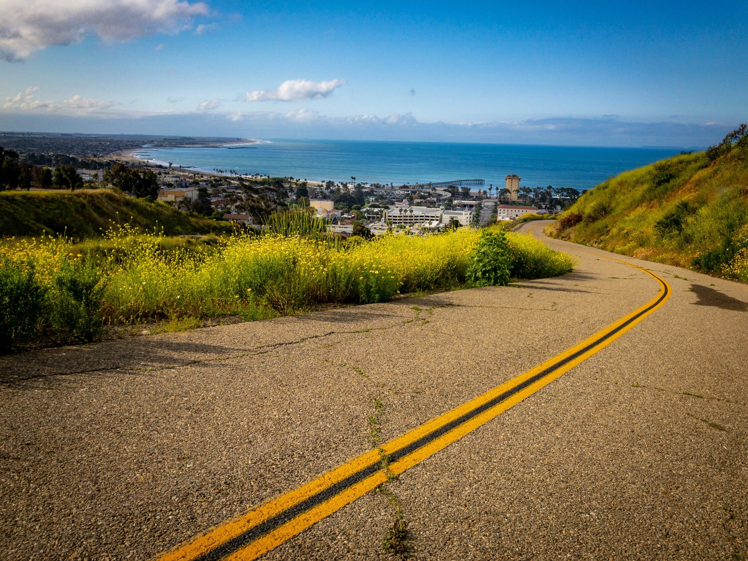 View of a coastal cityscape from a hillside road with yellow lane markings, overlooking the ocean with a pier and beach, under a partly cloudy sky.