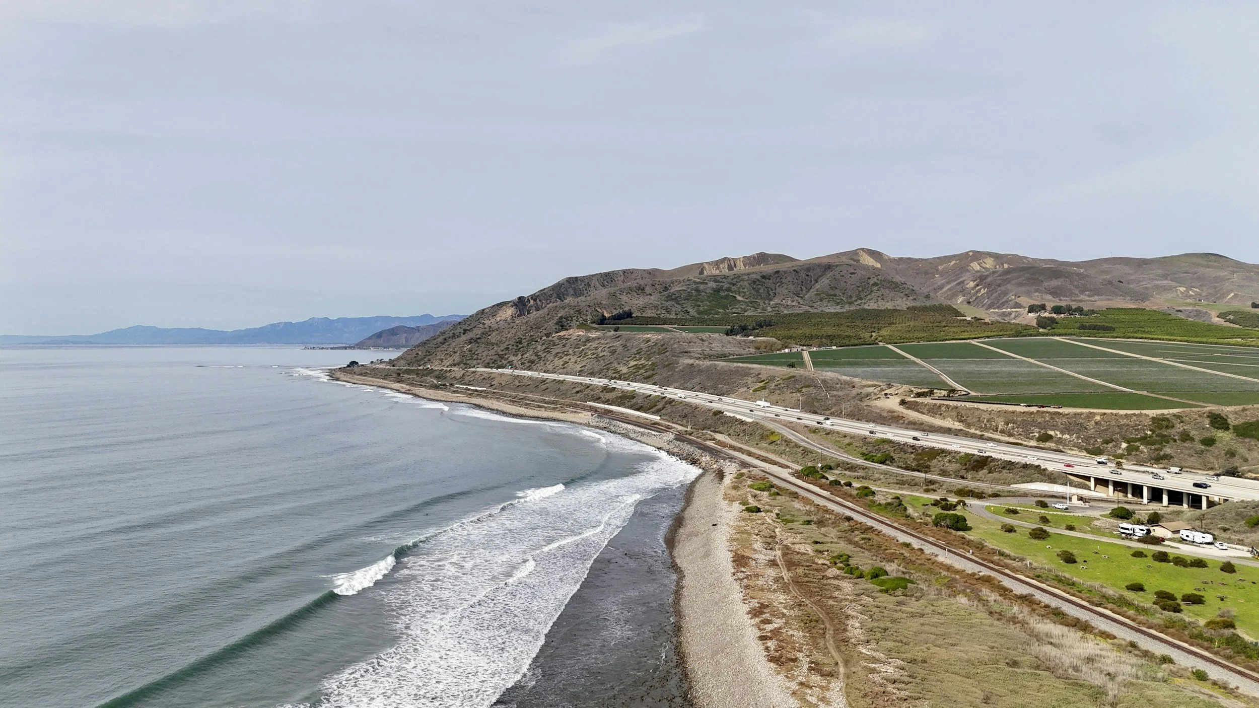 Coastal view with the ocean, shoreline, and rolling hills with green fields and a highway.