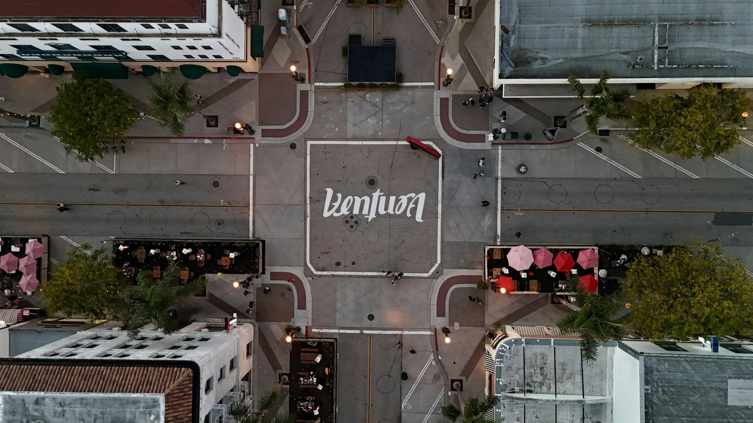 An aerial view of a downtown street intersection with the word 'Ventura' painted on the pavement. There are buildings, trees, streetlights, and a few pedestrians visible.