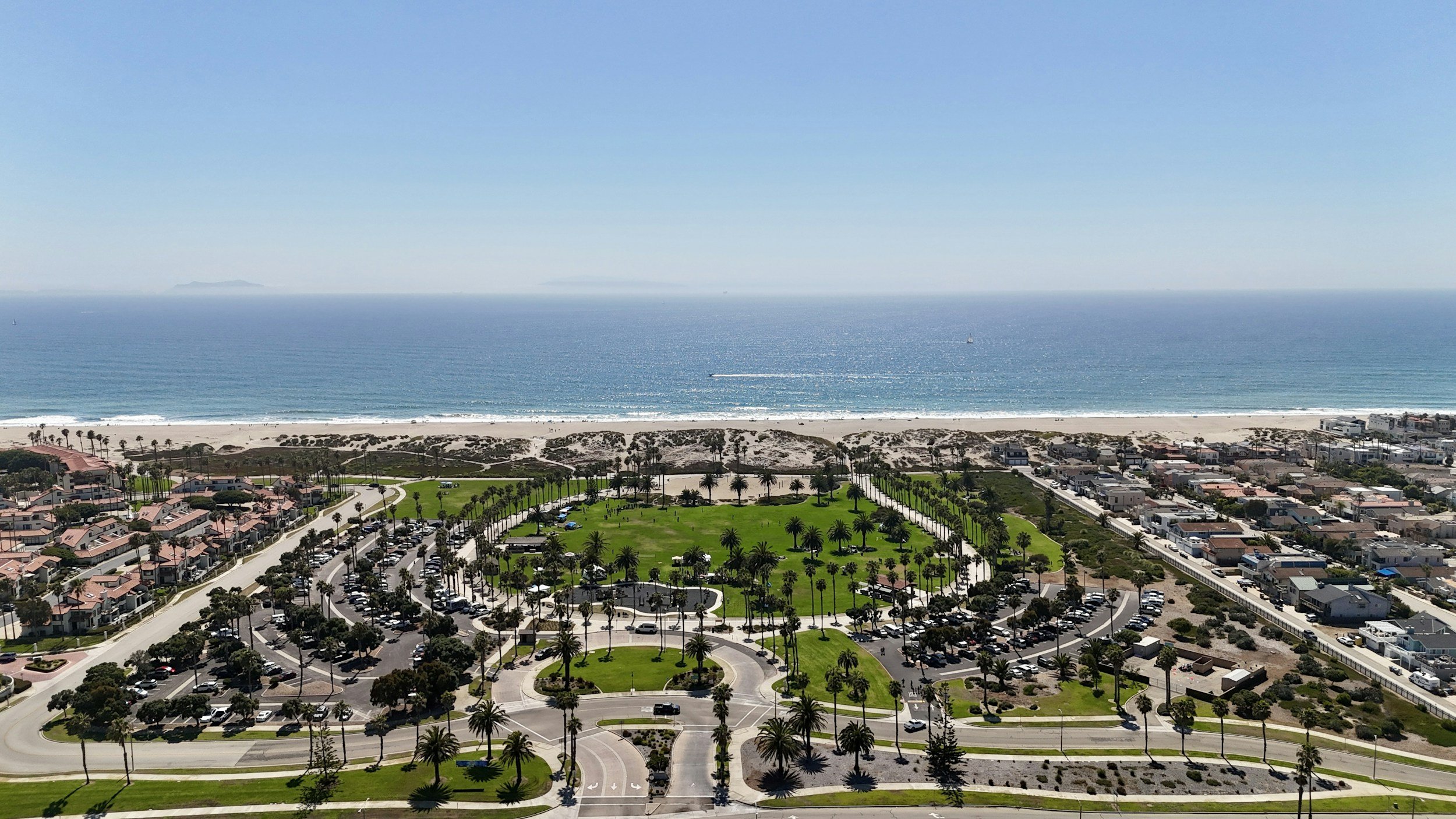 Aerial view of a coastal area with a beach, ocean, and residential neighborhoods with palm trees and a park.