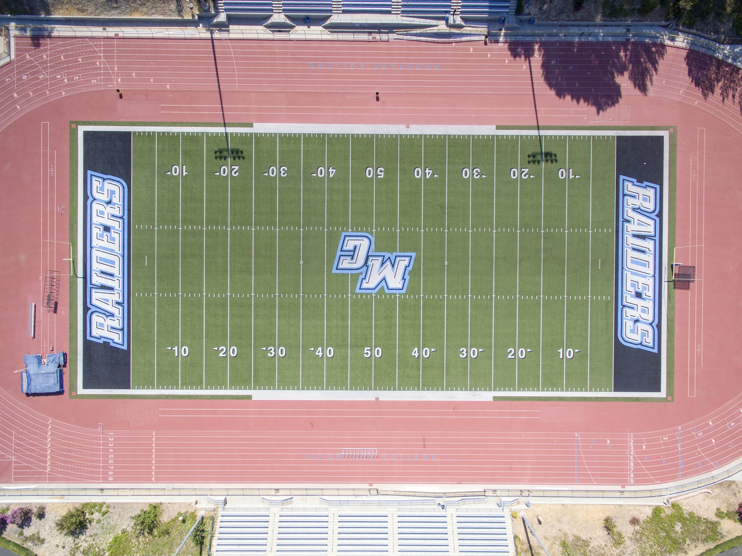 An aerial view of a college football field with green turf, large white and blue college logo at the center, and a red running track surrounding it.