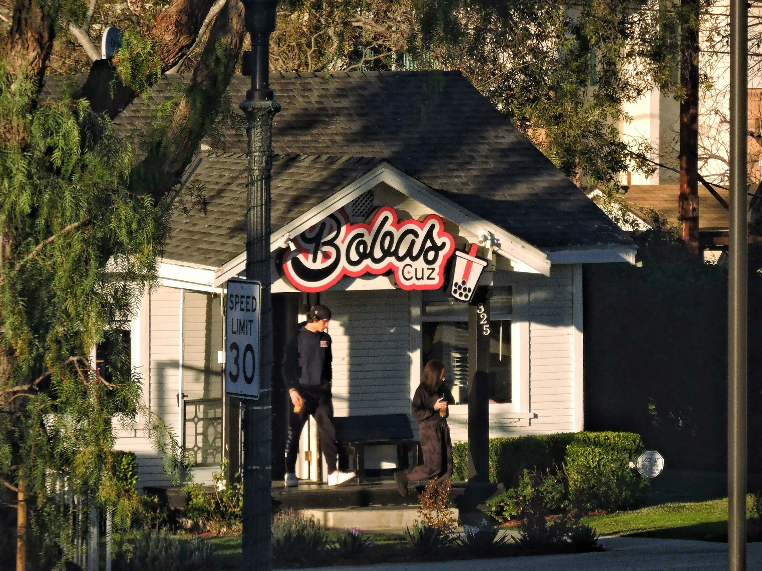 A small structure with a sign that reads 'Boba Cuz' with a bubble tea cup graphic. There are trees and electrical poles around, and two people are walking in front of the building.